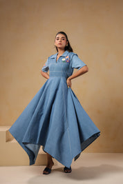 Model wearing the Daisydrift Handkerchief Dress in soft denim, showcasing a dramatic handkerchief hem and structured fit with floral embroidery on the chest.