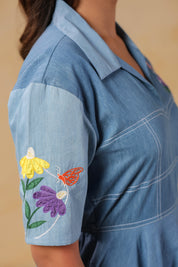 Macro shot of the sleeve embroidery showing yellow, purple, and orange floral motifs with butterfly detail on a soft denim canvas.