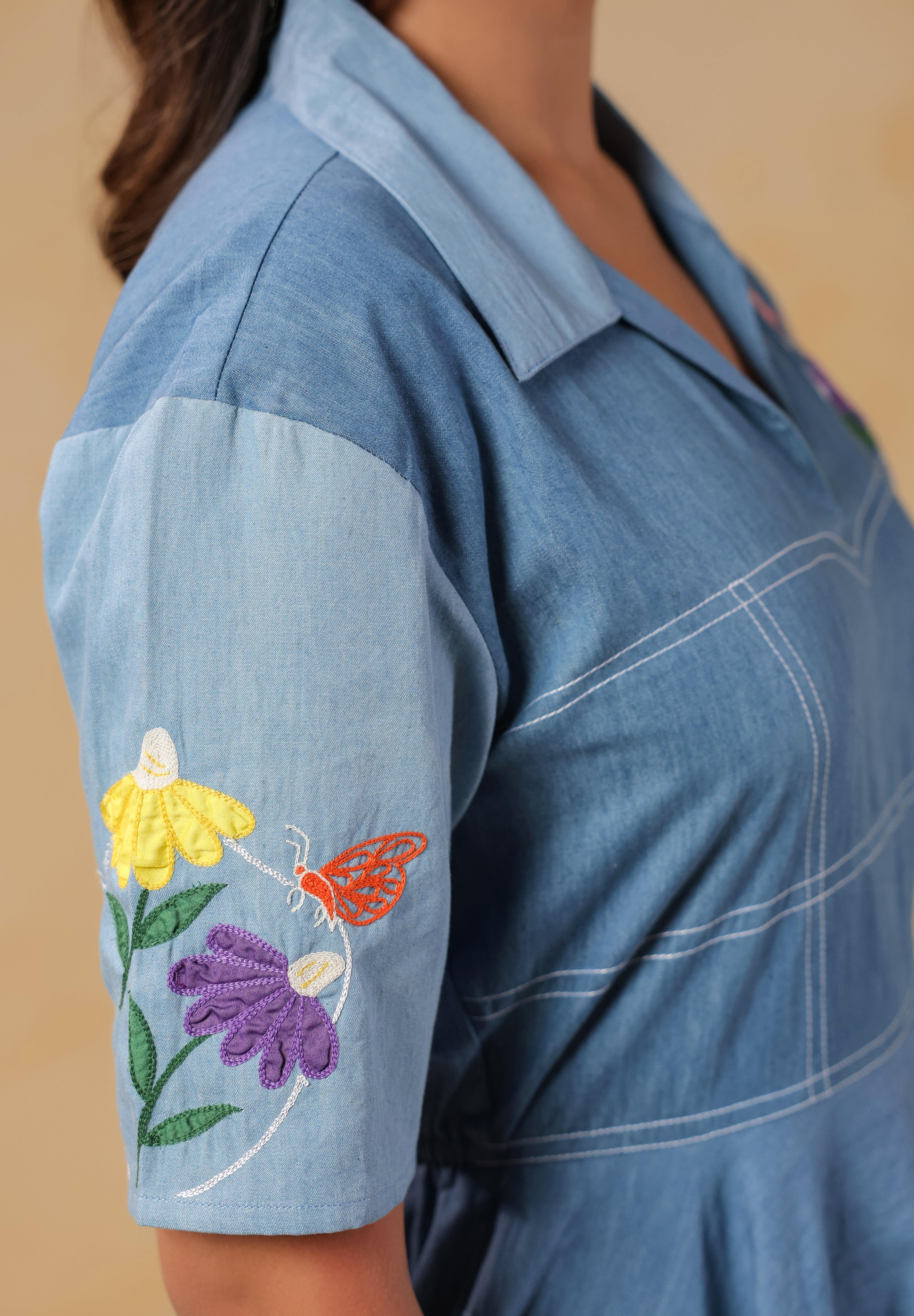 Macro shot of the sleeve embroidery showing yellow, purple, and orange floral motifs with butterfly detail on a soft denim canvas.