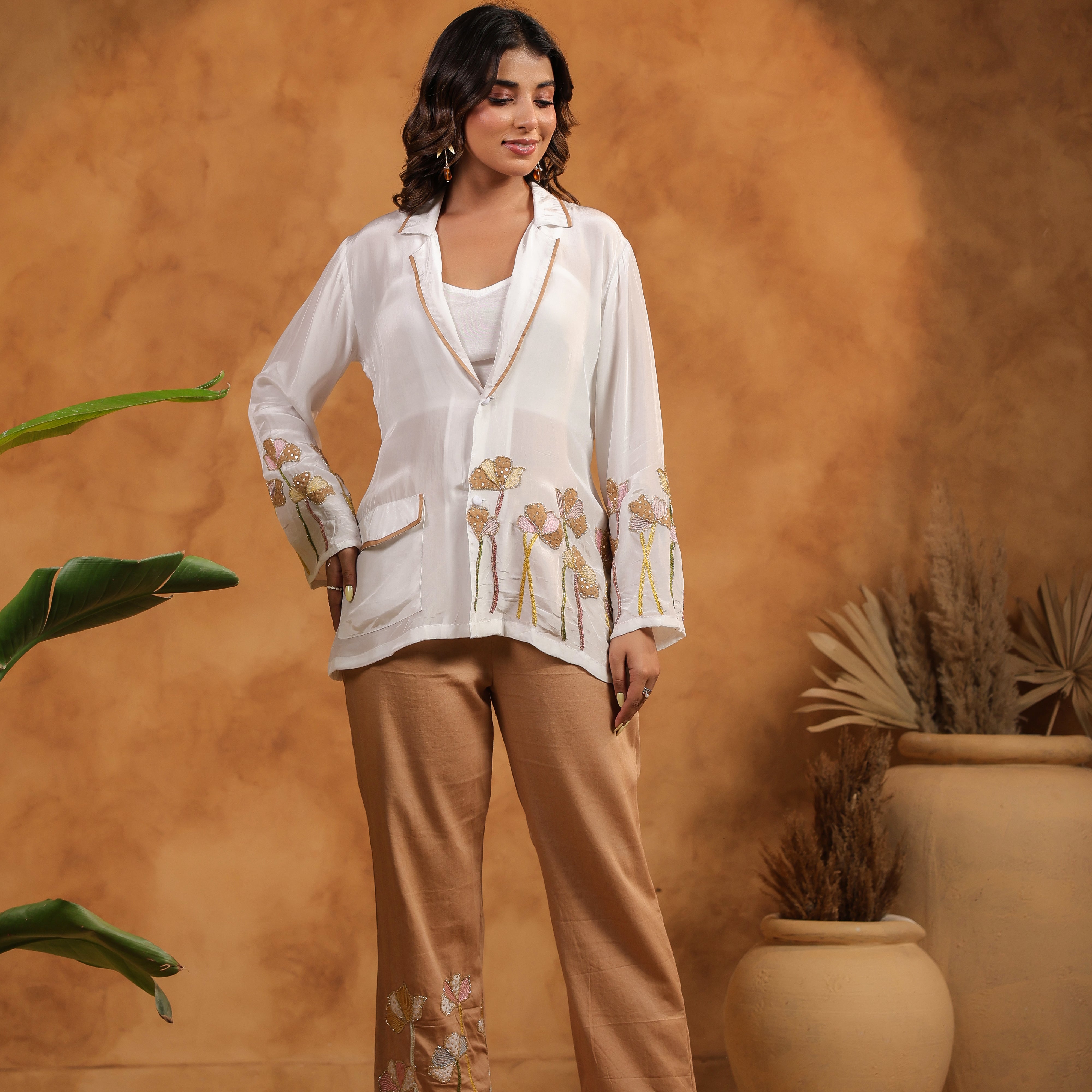 Woman in a white blouse and beige pants standing against an orange wall with plants.