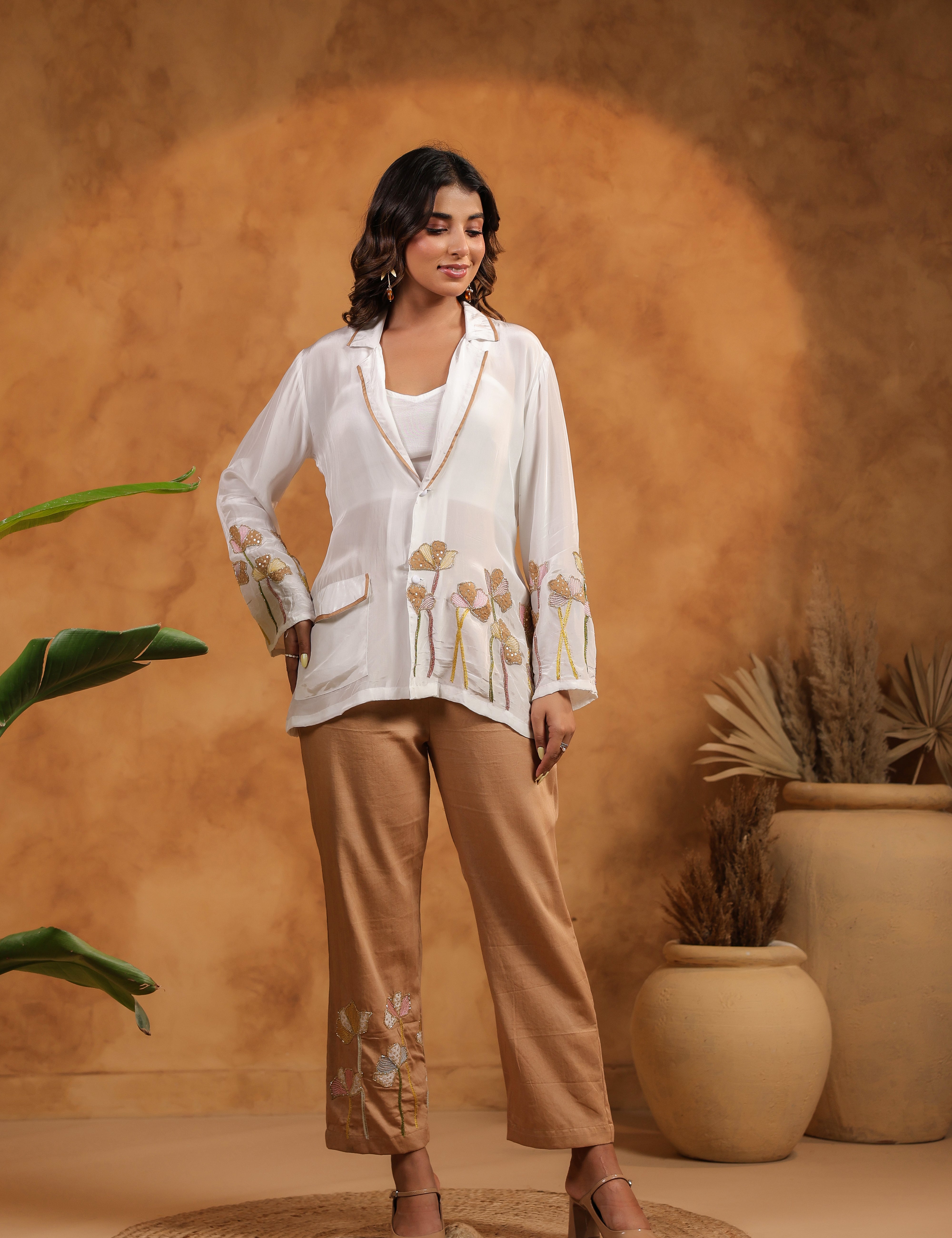 Woman in a white blouse and beige pants standing against an orange wall with plants.