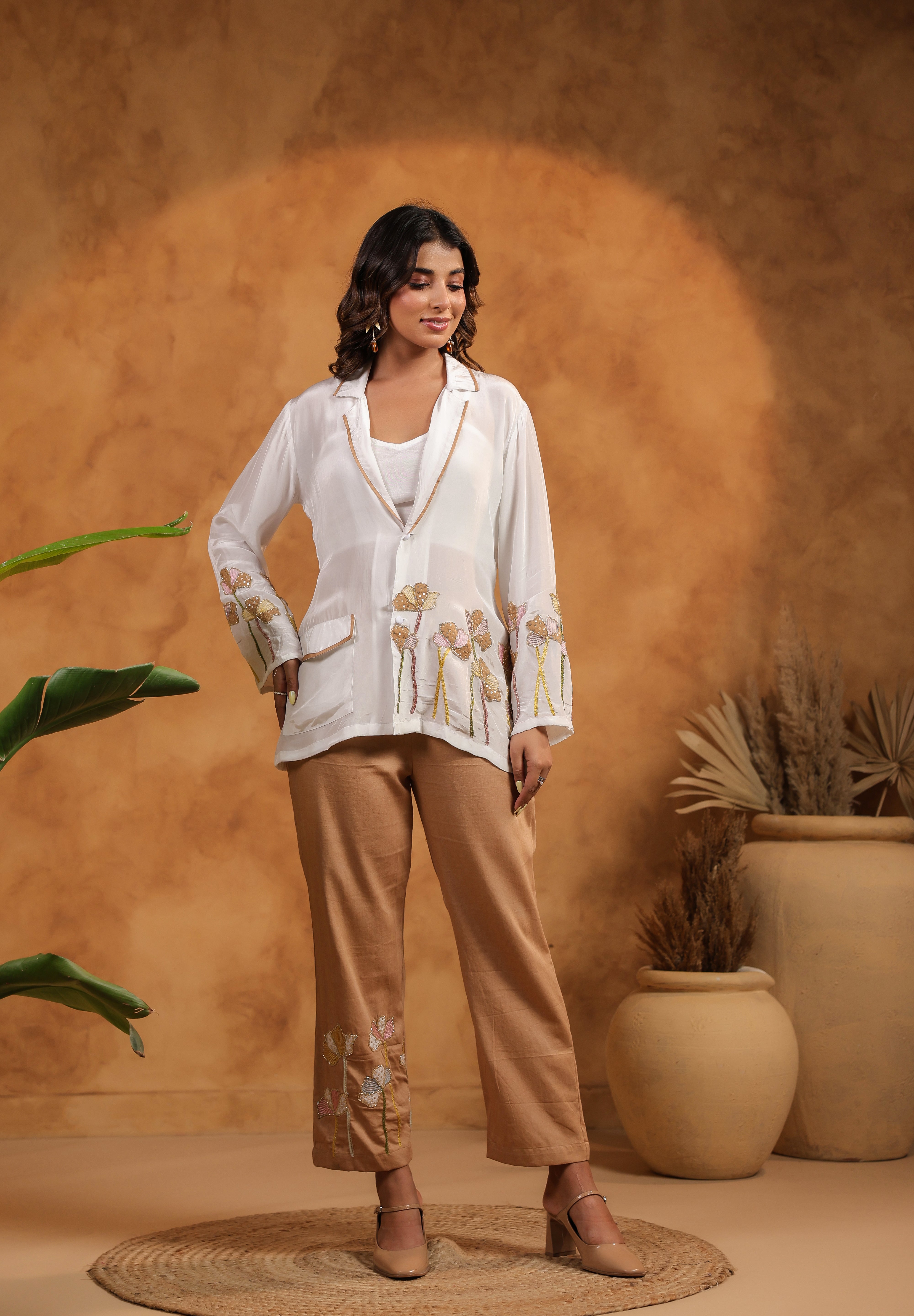 Woman in a white blouse and beige pants standing against an orange wall with plants.