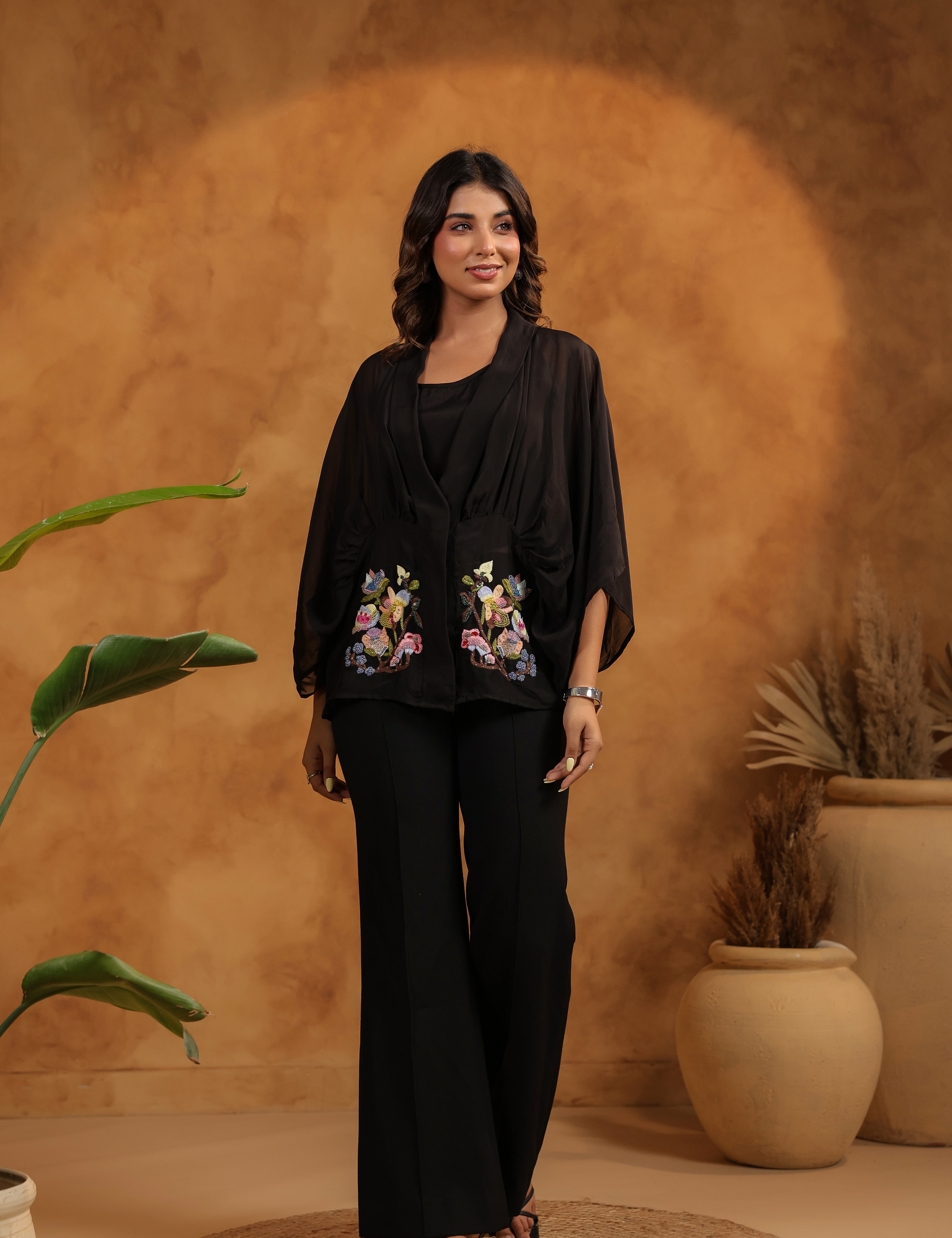 Woman in black outfit with floral embroidery standing against a warm-toned wall with plants.