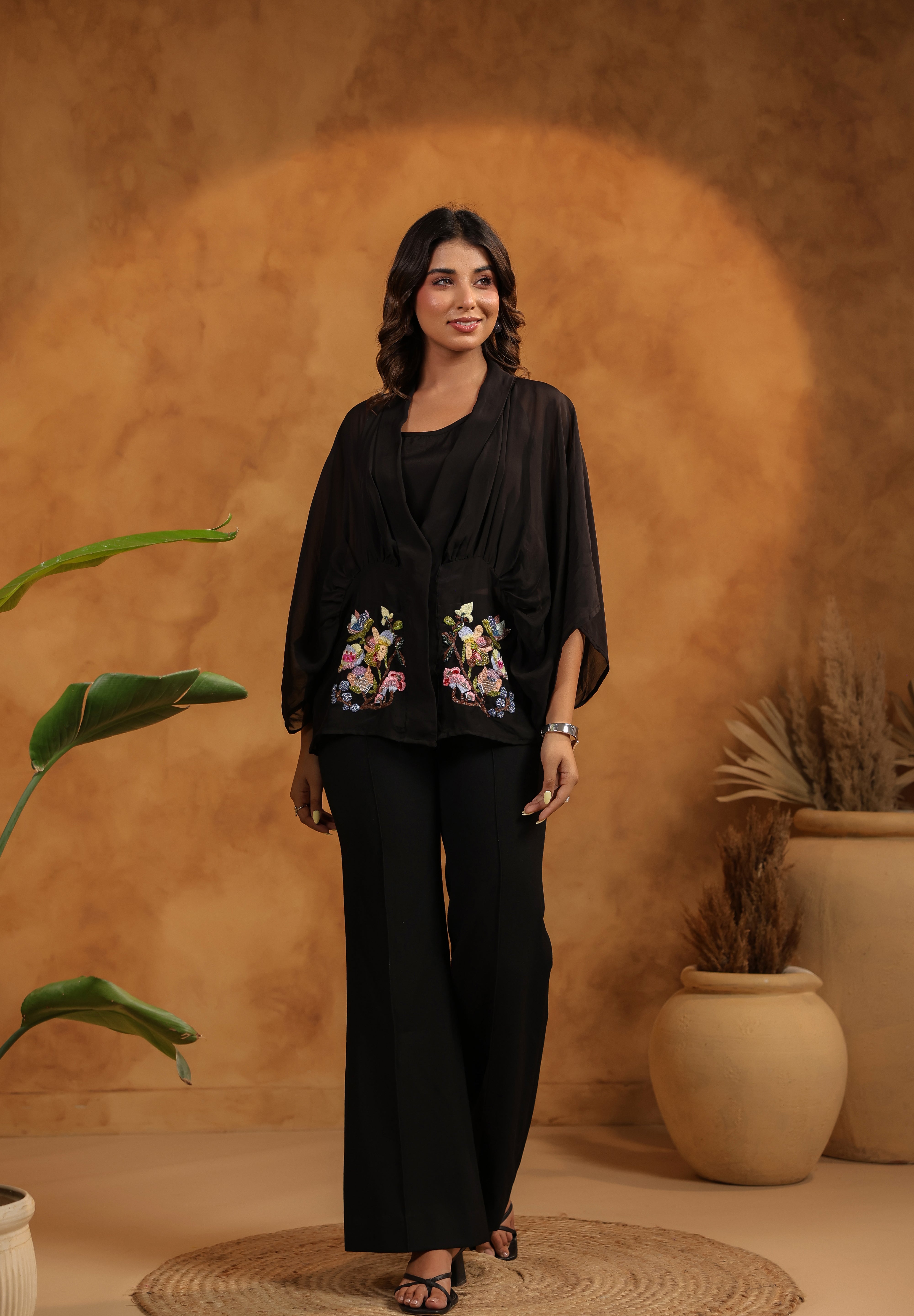 Woman in black outfit with floral embroidery standing against a warm-toned wall with plants.