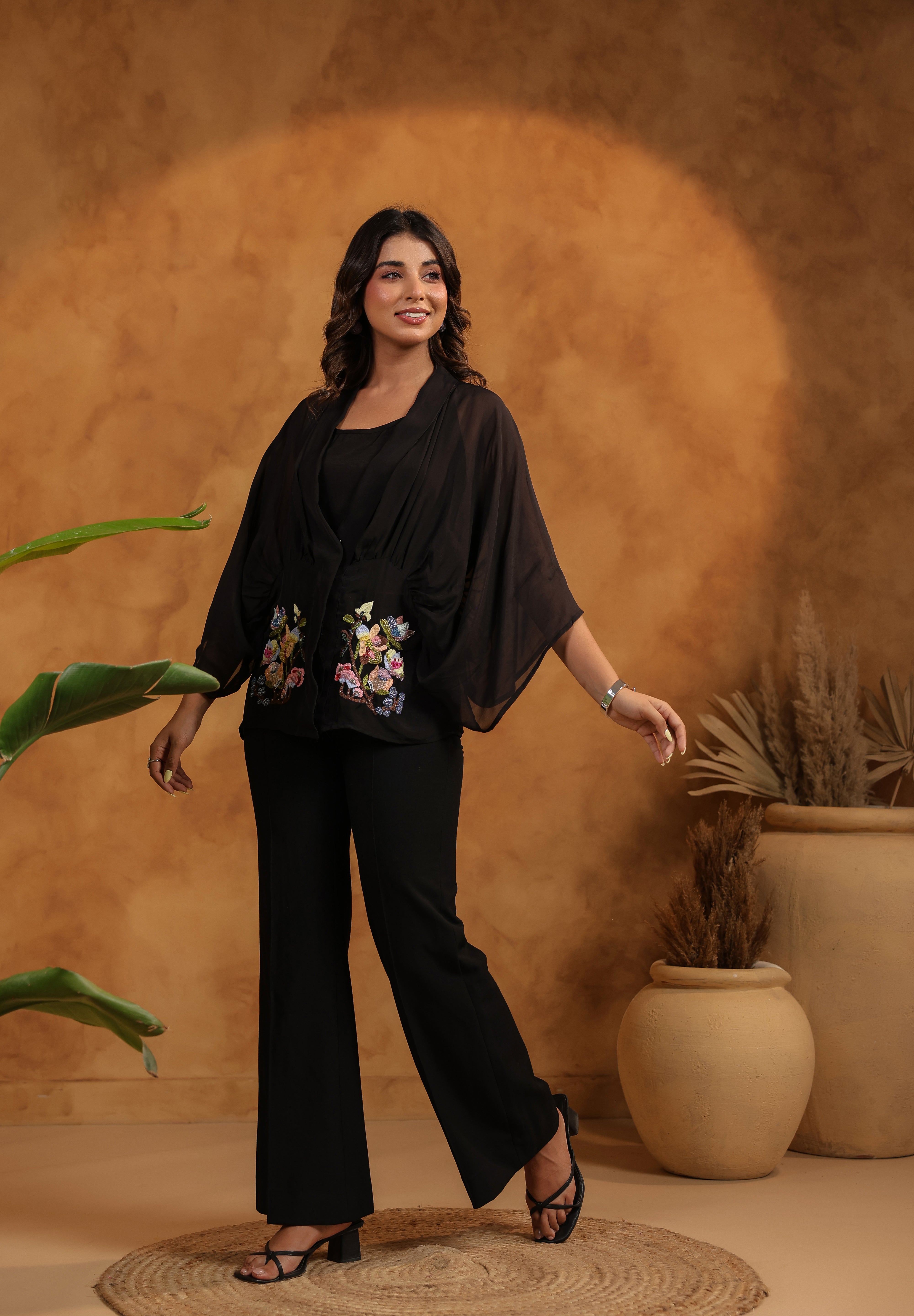 Woman in a black outfit with floral embroidery standing against a warm-toned wall with plants.