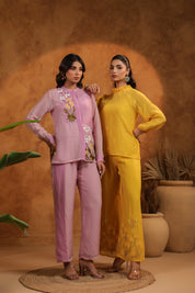 Two women in pink and yellow outfits standing against a warm-toned background with plants.