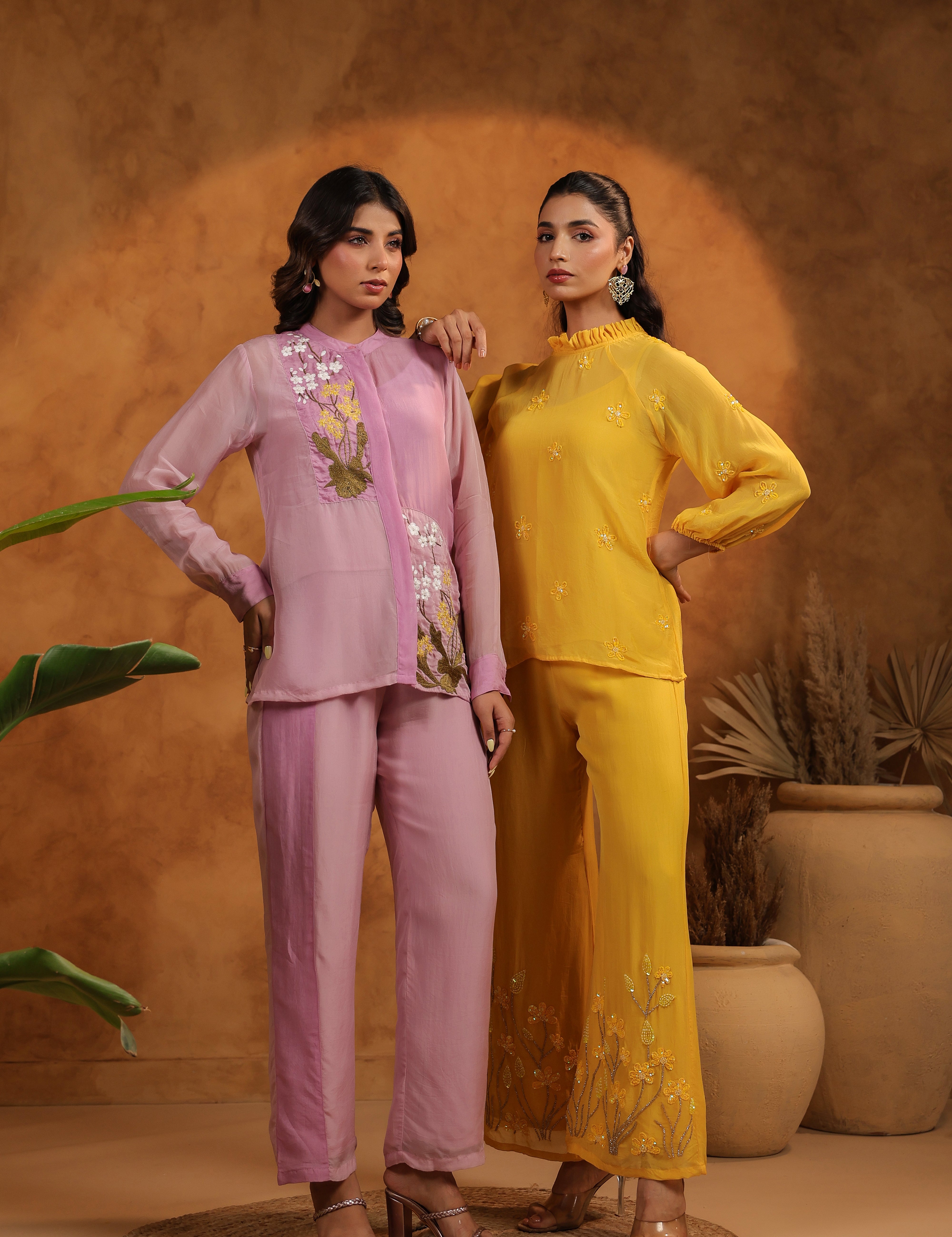 Two women in pink and yellow outfits standing against a warm-toned background with plants.
