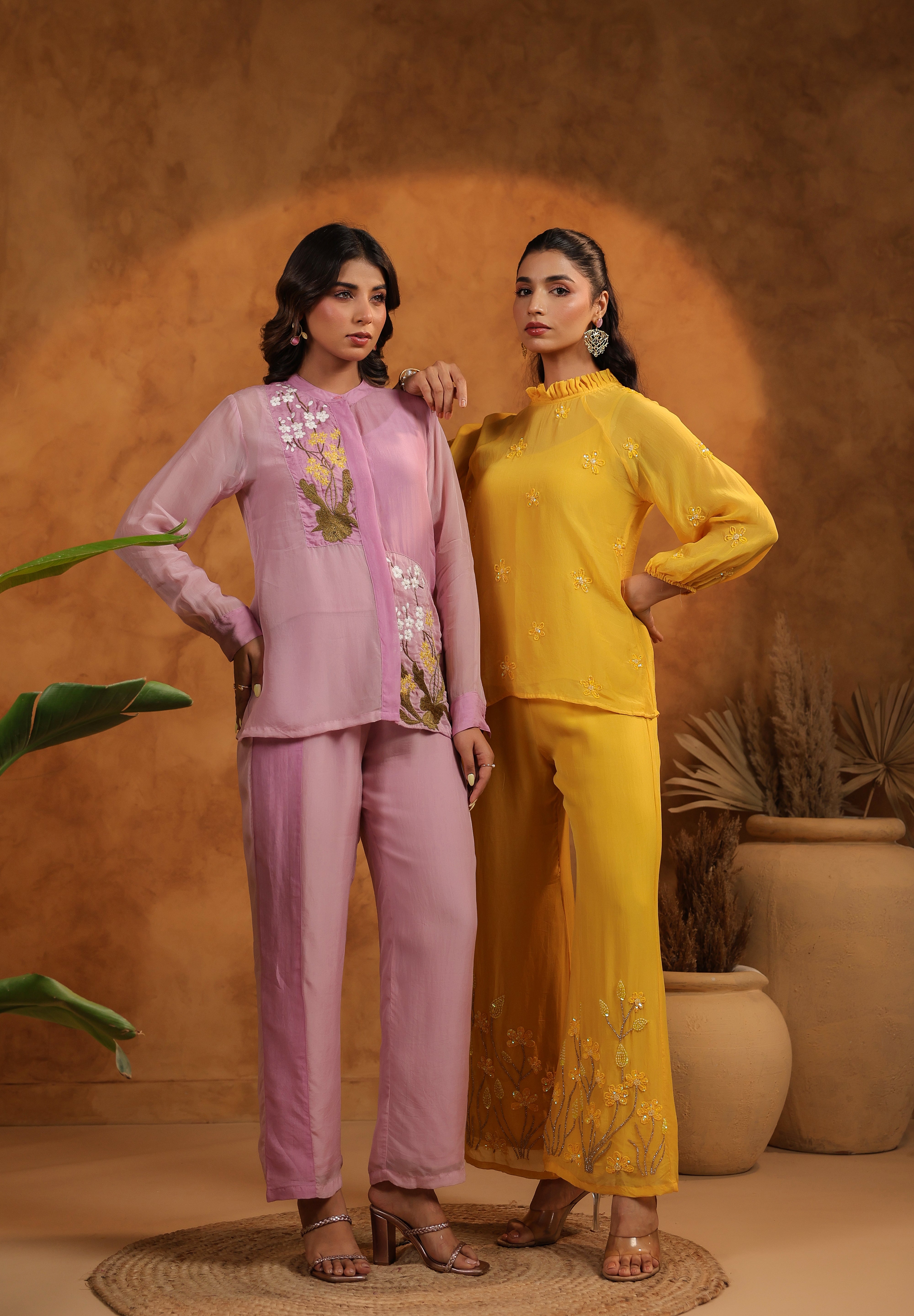 Two women in pink and yellow outfits standing against a warm-toned background with plants.