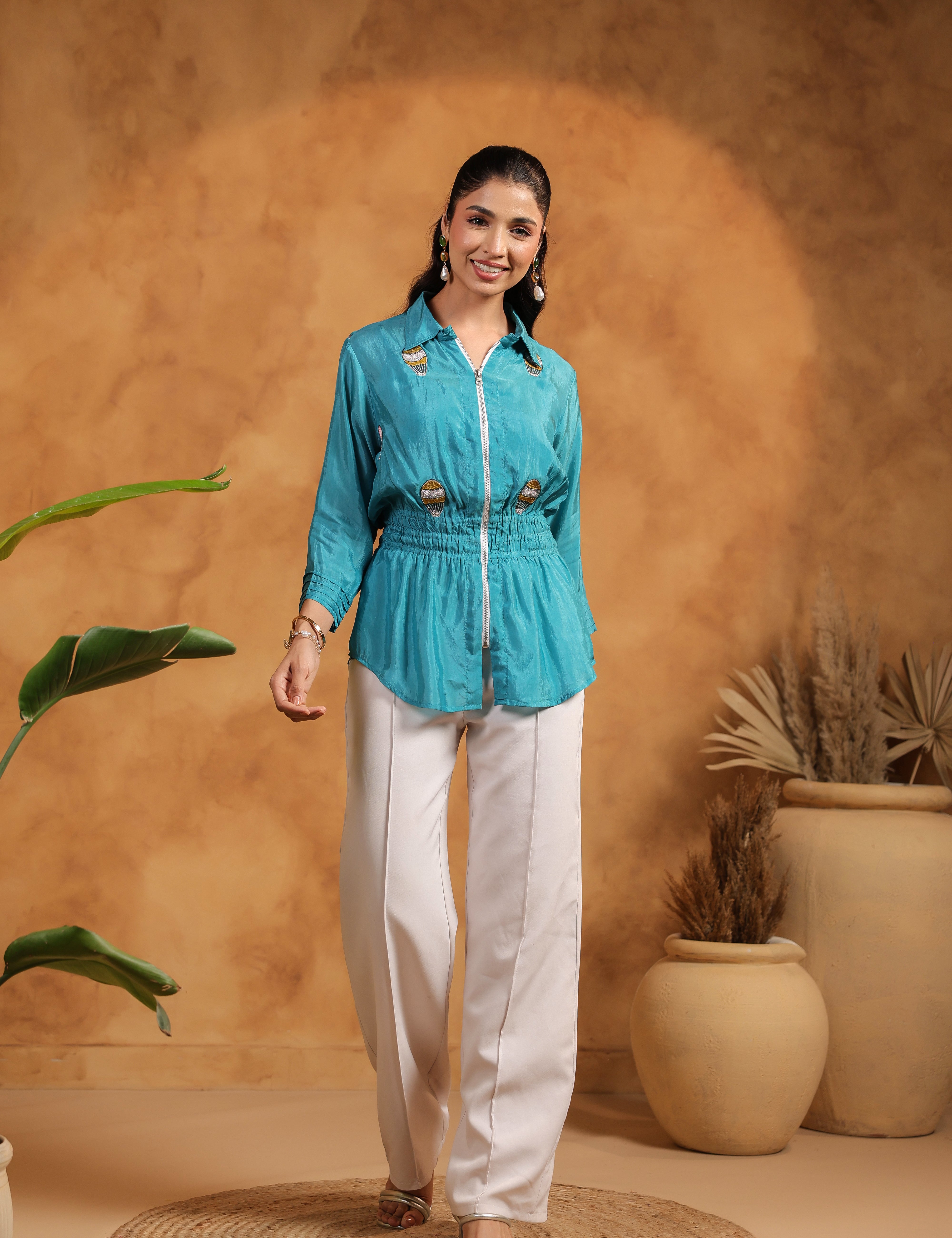 Woman in a turquoise top and white pants standing against a warm-toned wall with plants.