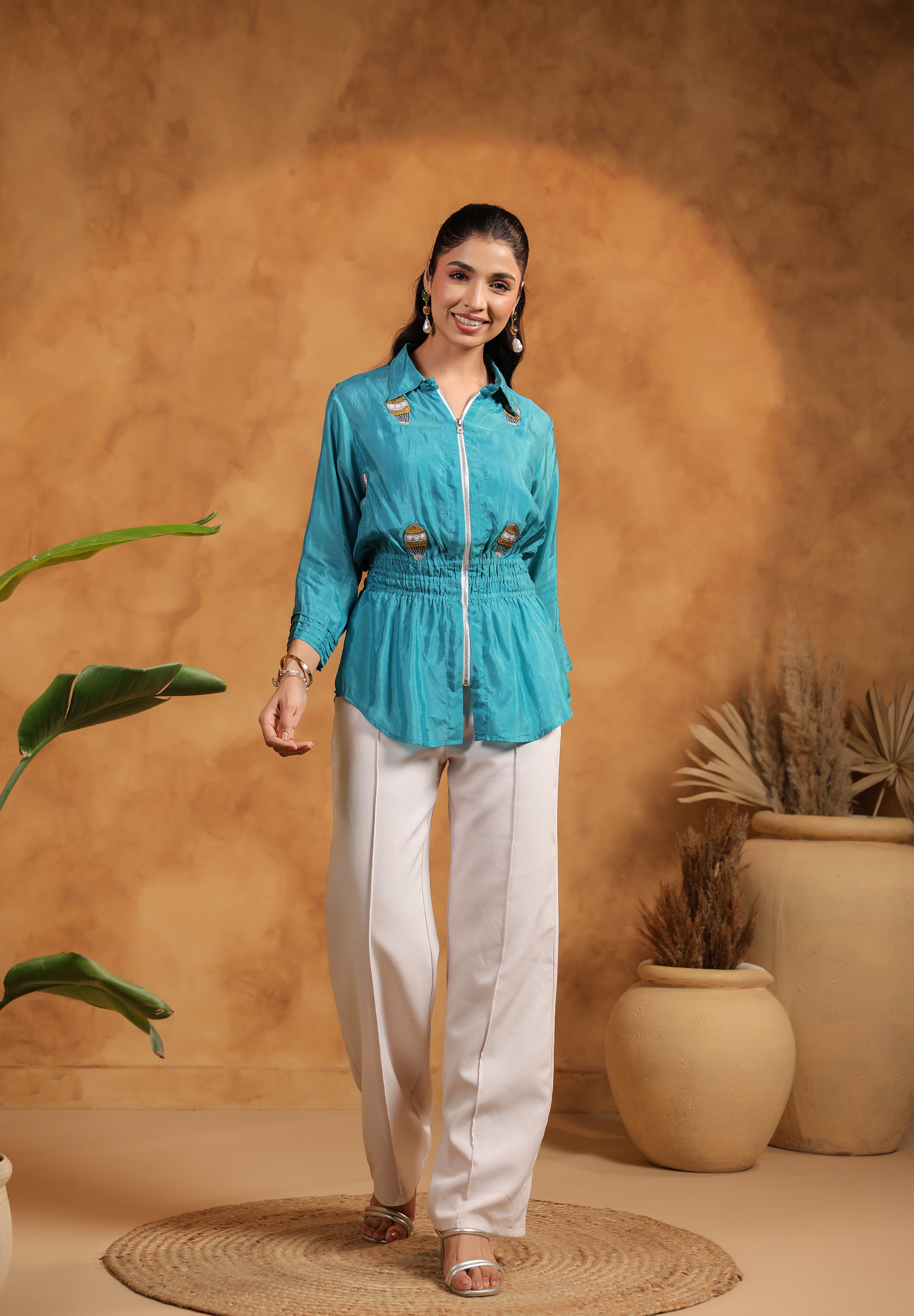 Woman in a turquoise top and white pants standing against a warm-toned wall with plants.