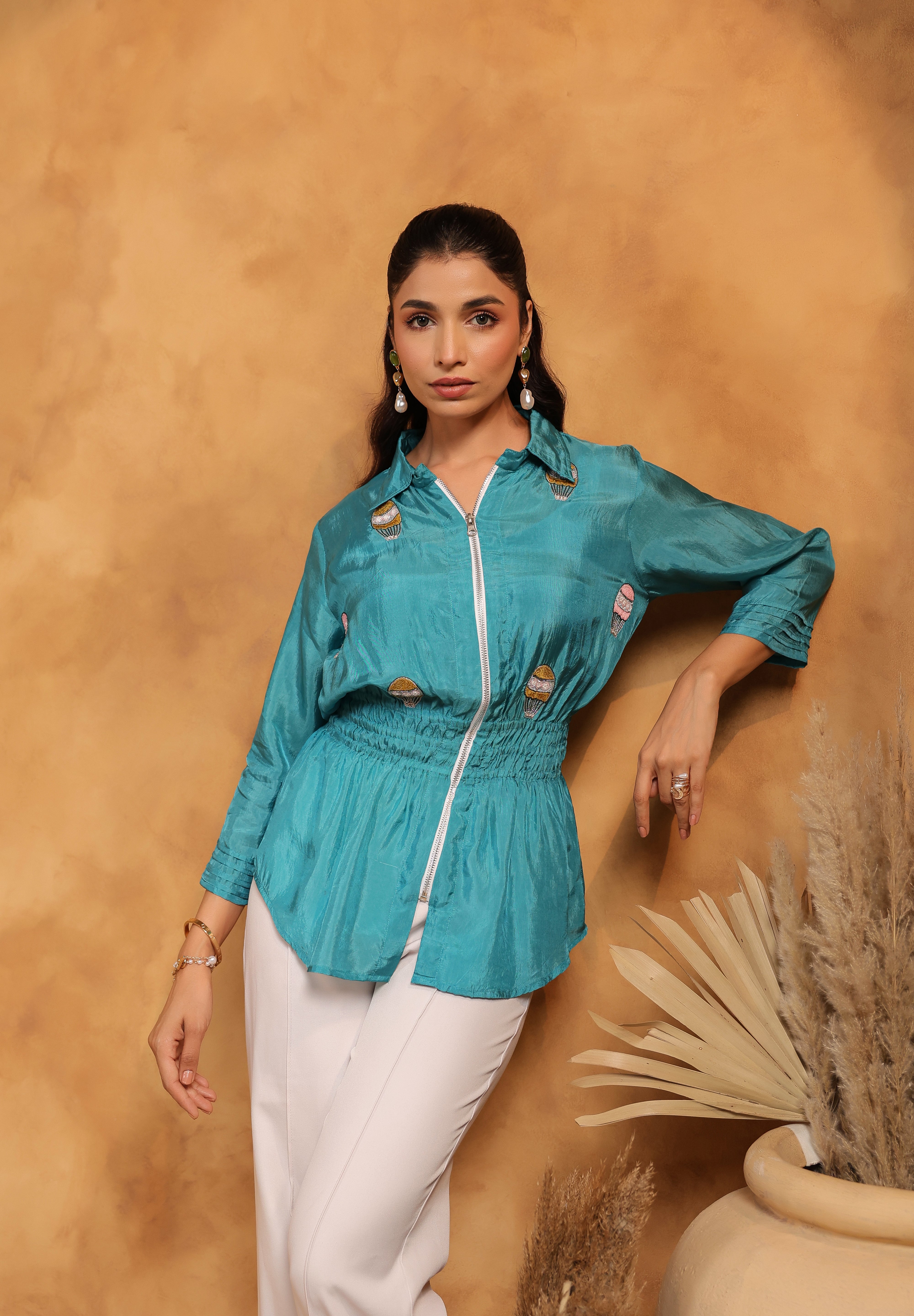 Woman wearing a teal jacket with white pants against a beige background
