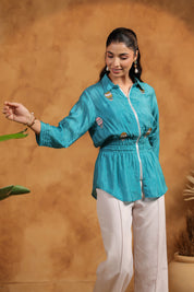 Woman wearing a turquoise shirt and white pants against a brown background