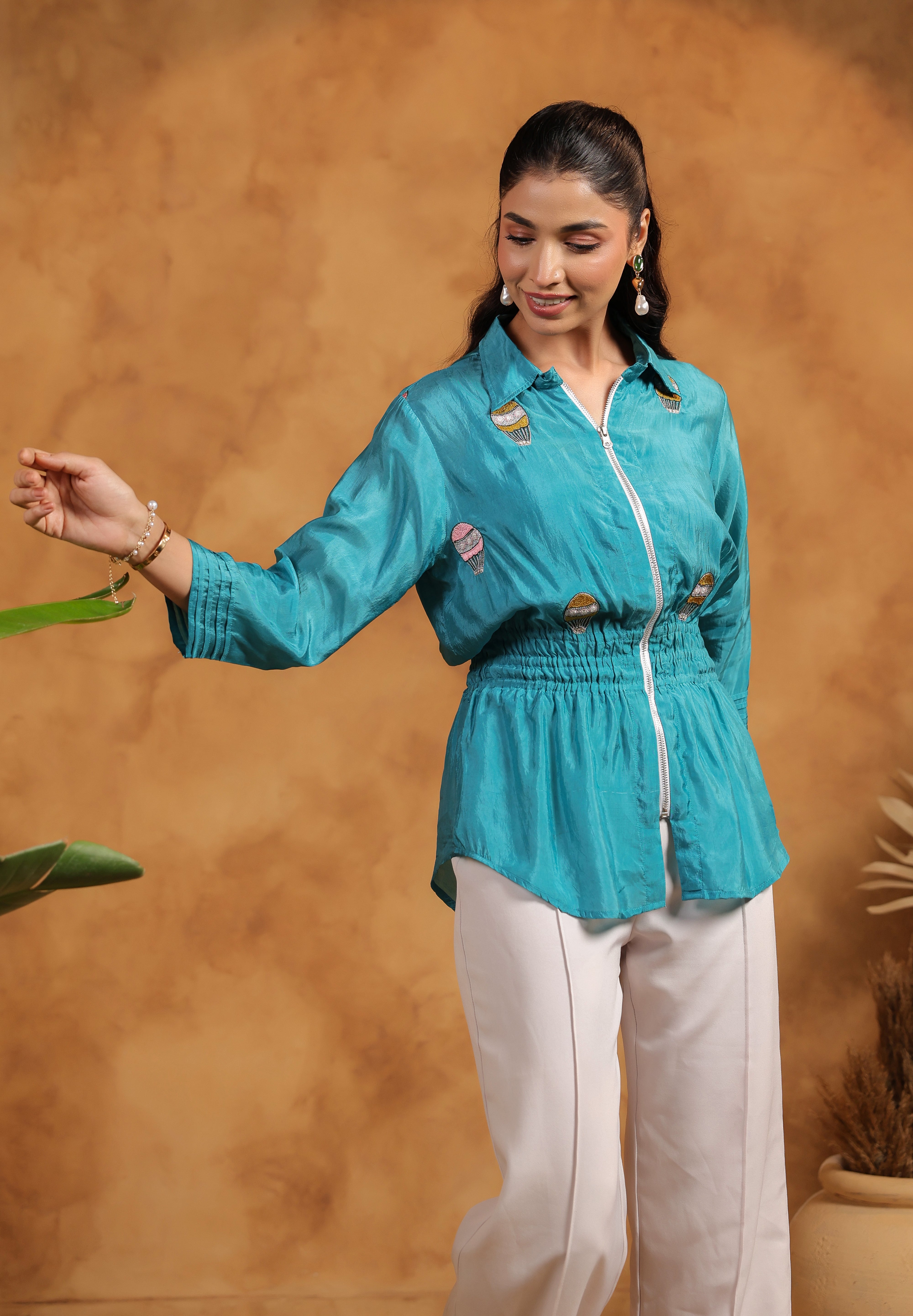 Woman wearing a turquoise shirt and white pants against a brown background