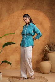 Woman wearing a turquoise shirt and white pants against a warm-toned wall with plants.