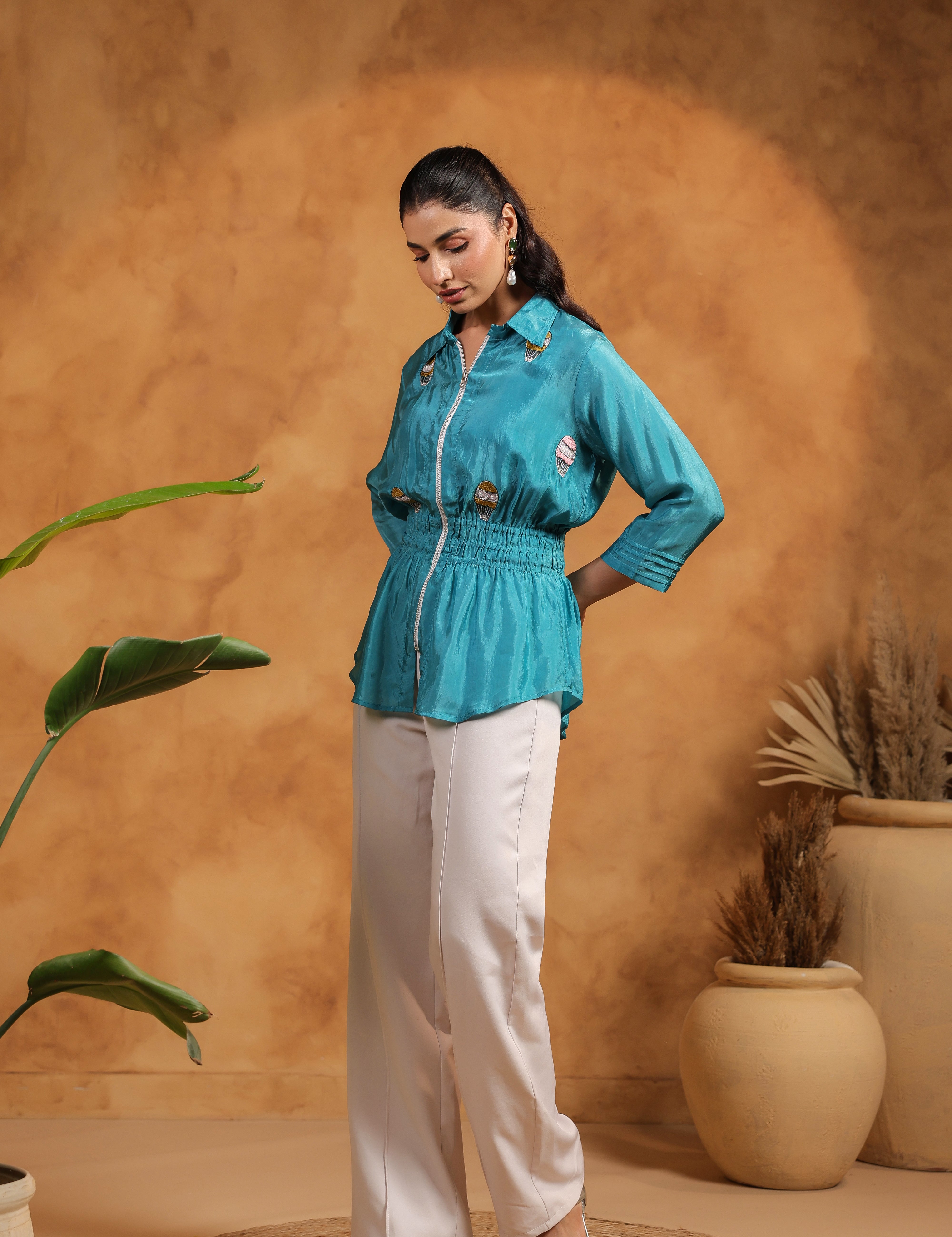 Woman wearing a turquoise shirt and white pants against a warm-toned wall with plants.