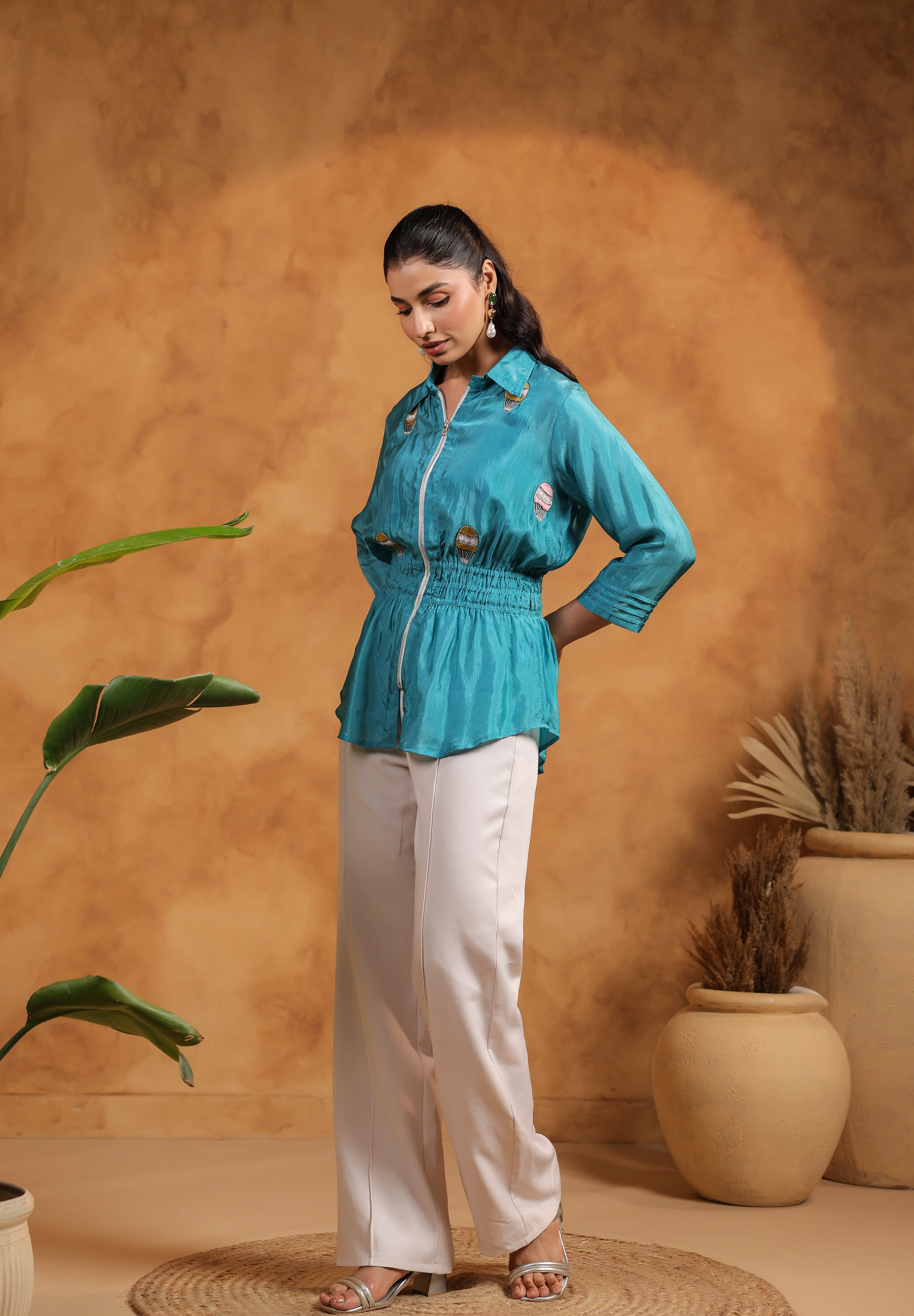 Woman wearing a turquoise shirt and white pants against a warm-toned wall with plants.