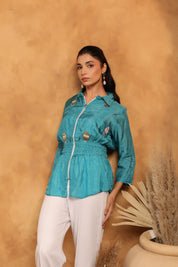 Woman wearing a teal shirt with white pants against a beige wall.
