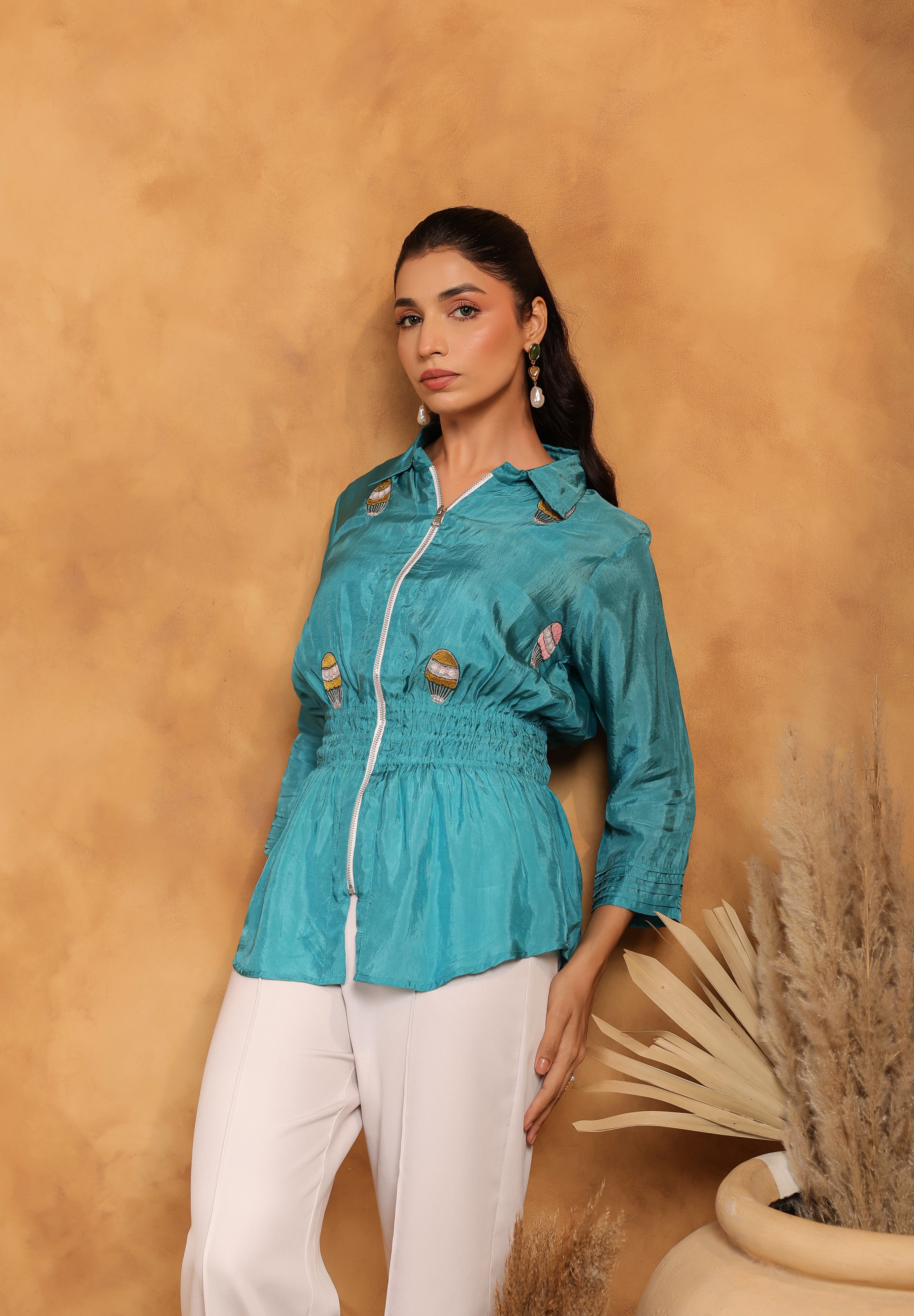 Woman wearing a teal shirt with white pants against a beige wall.