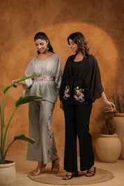 Two women in stylish outfits standing in a room with a textured wall and potted plants.