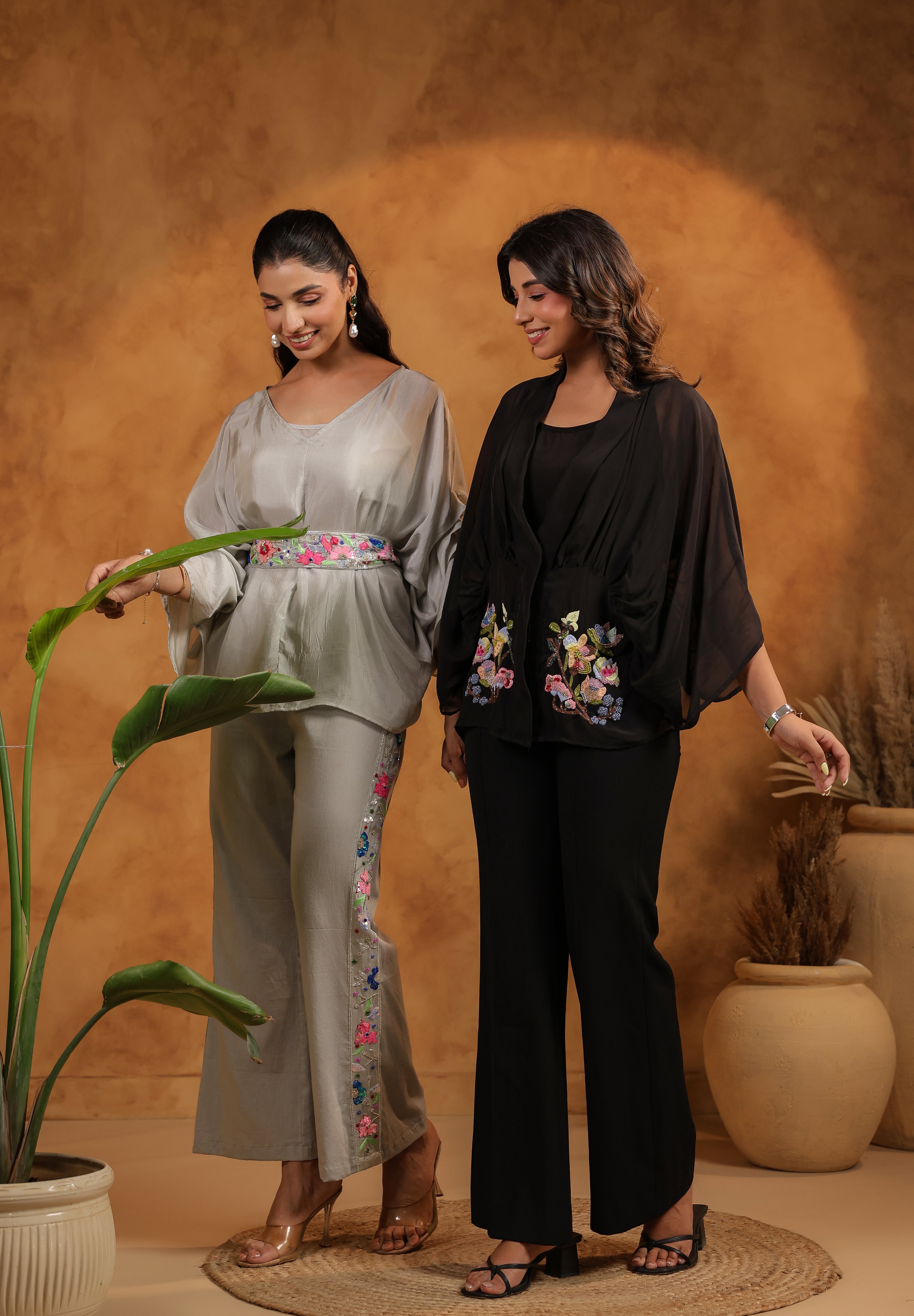 Two women in stylish outfits standing in a room with a textured wall and potted plants.