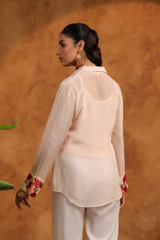 Woman wearing a light pink blouse with floral cuffs and white pants against an orange background