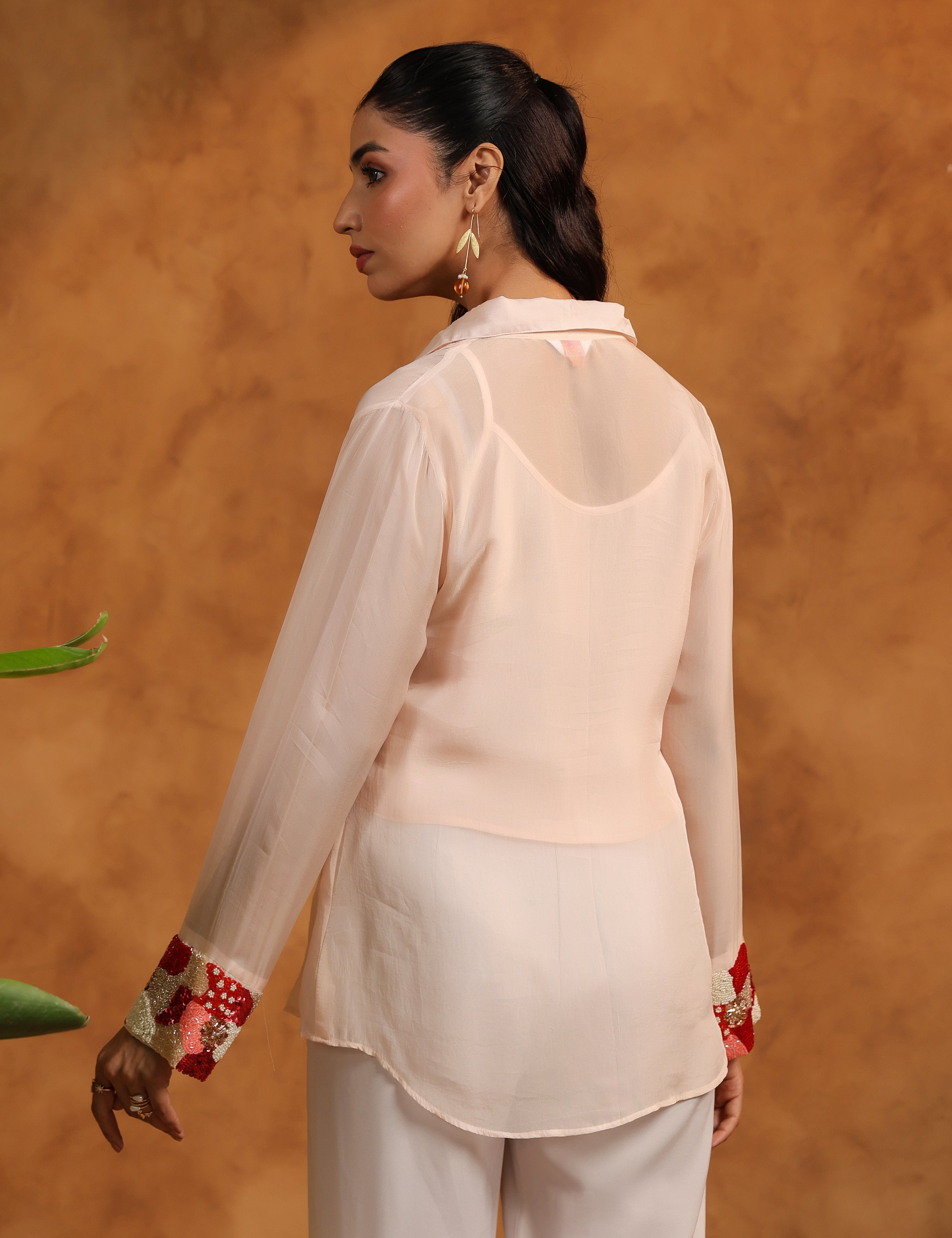 Woman wearing a light pink blouse with floral cuffs and white pants against an orange background