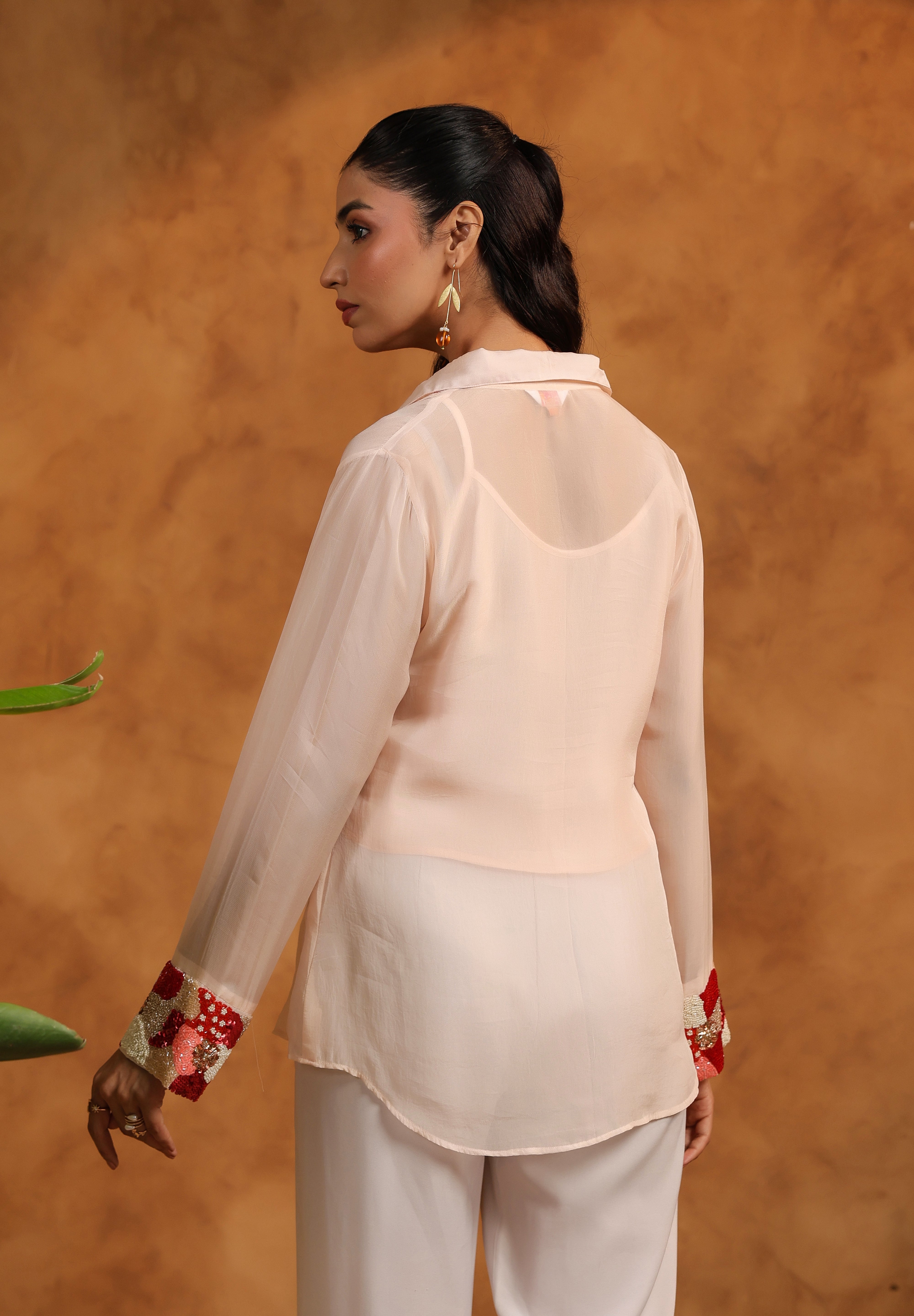 Woman wearing a light pink blouse with floral cuffs and white pants against an orange background