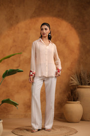 Woman wearing a light-colored blouse and pants against a warm-toned wall with plants.