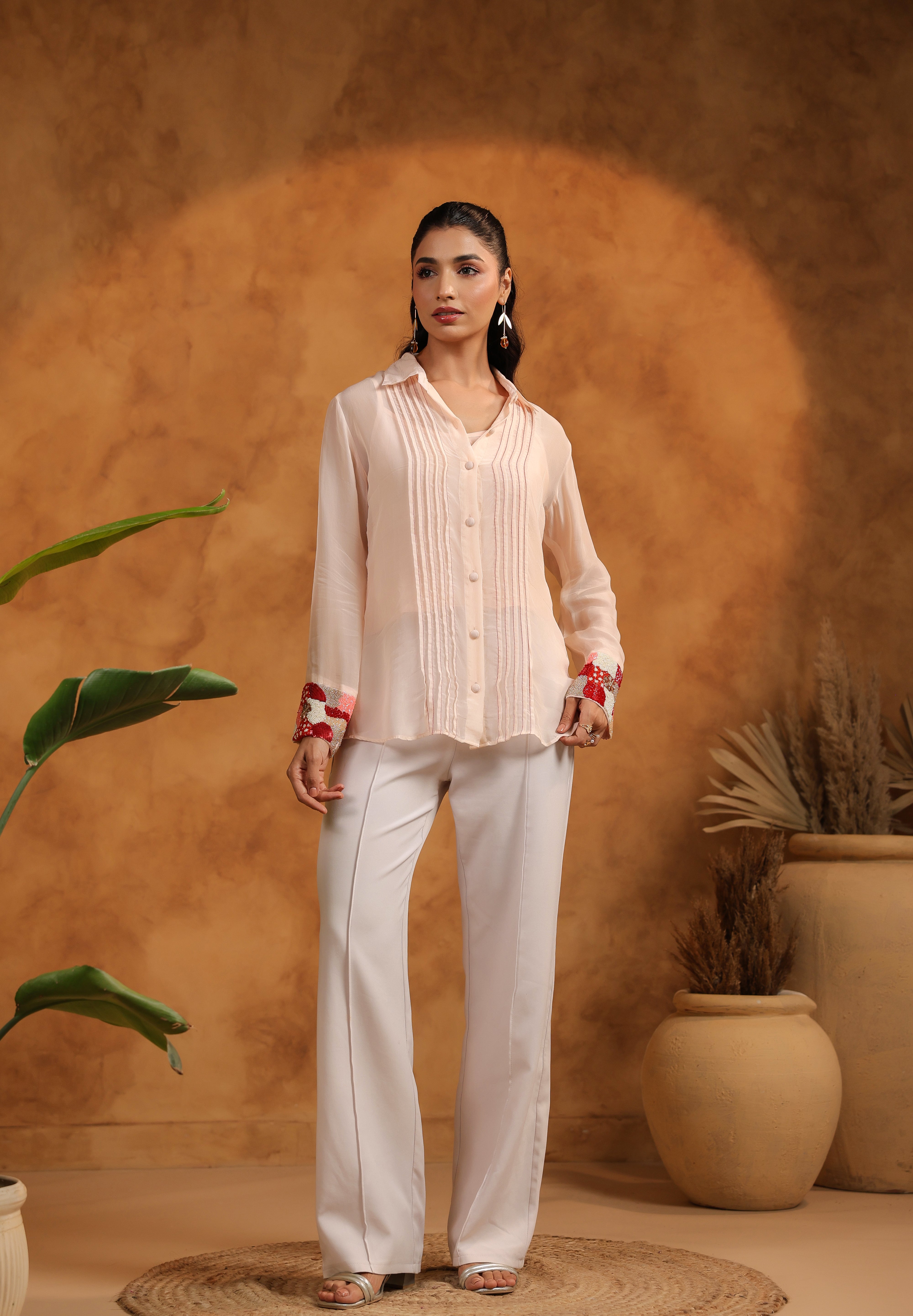 Woman wearing a light-colored blouse and pants against a warm-toned wall with plants.
