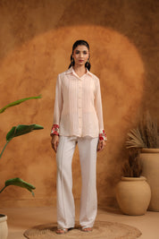 Woman in a pink blouse and white pants standing against an orange wall with plants.
