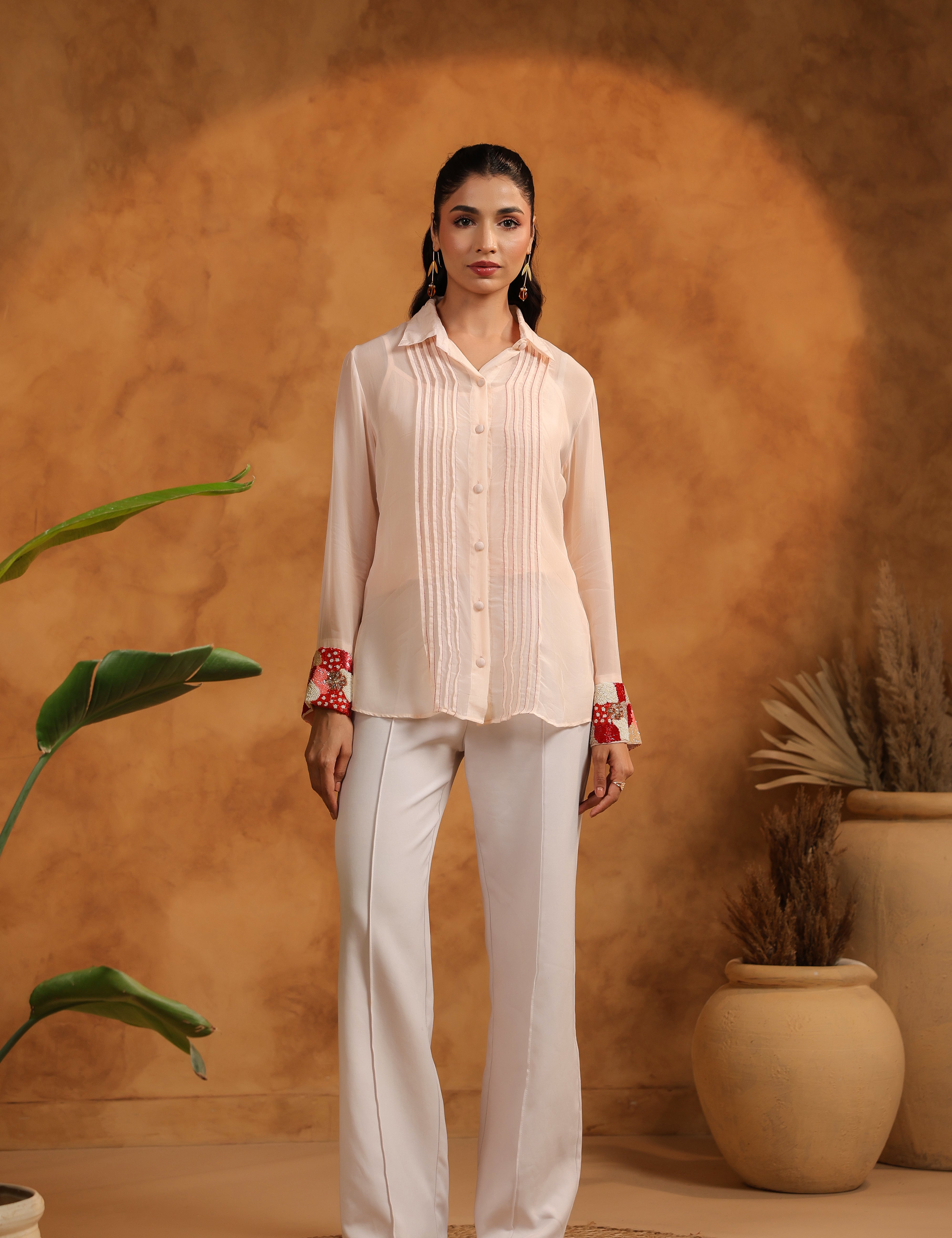 Woman in a pink blouse and white pants standing against an orange wall with plants.