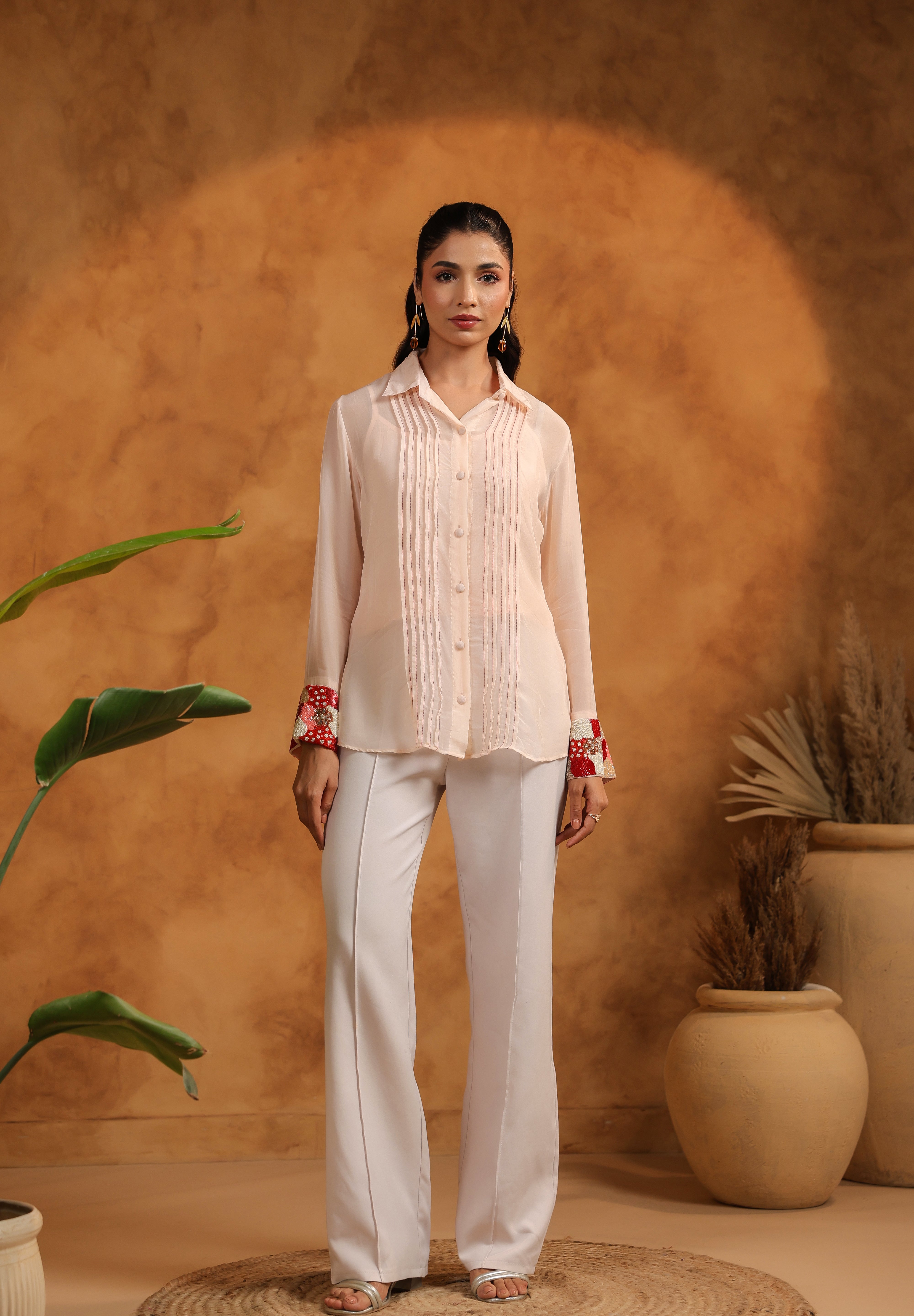 Woman in a pink blouse and white pants standing against an orange wall with plants.