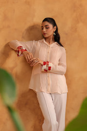 Woman wearing a light-colored blouse with red embellishment and white pants against a warm-toned wall.