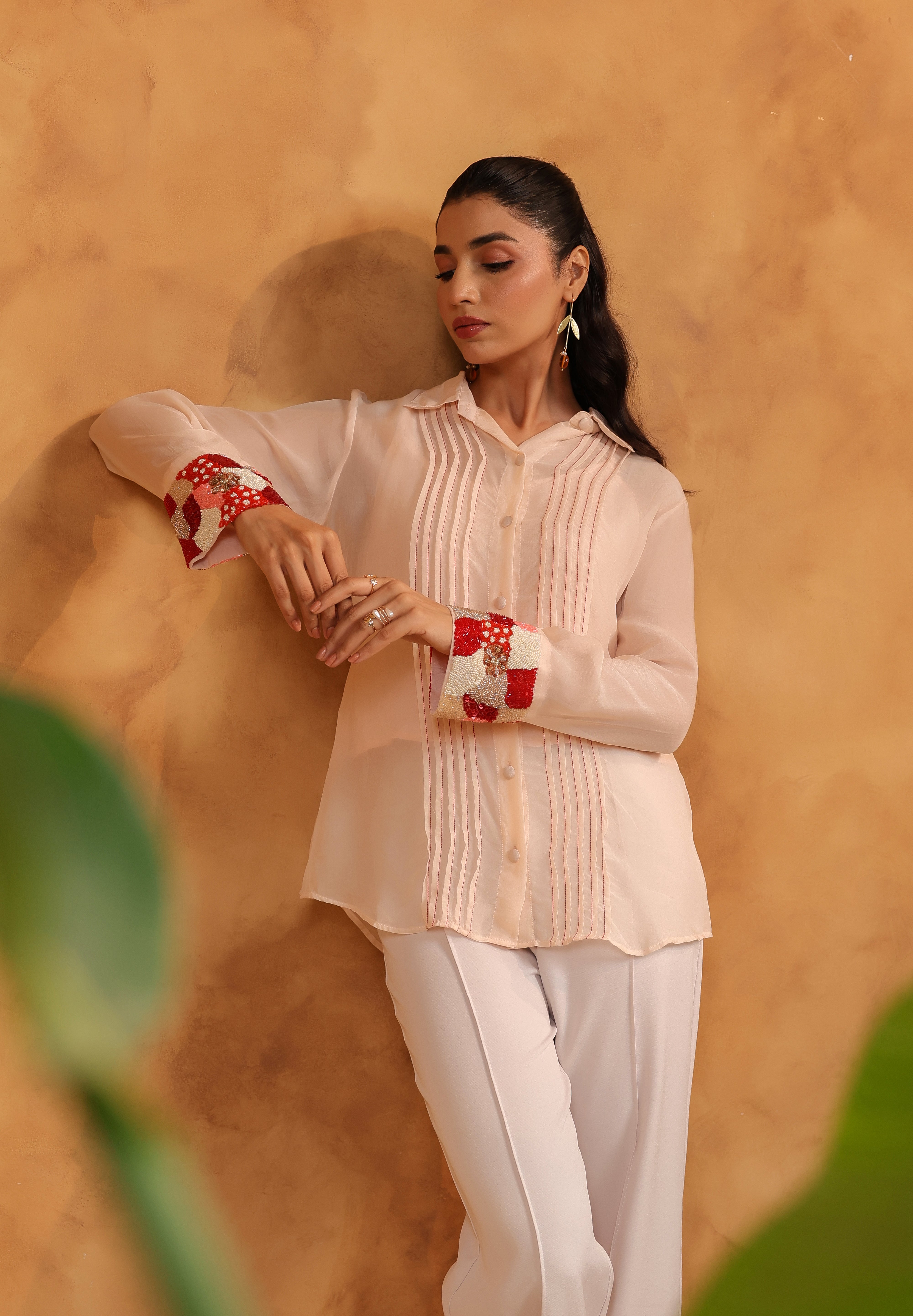 Woman wearing a light-colored blouse with red embellishment and white pants against a warm-toned wall.
