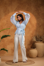 Woman in a light blue top and white pants posing against a warm brown background with plants.