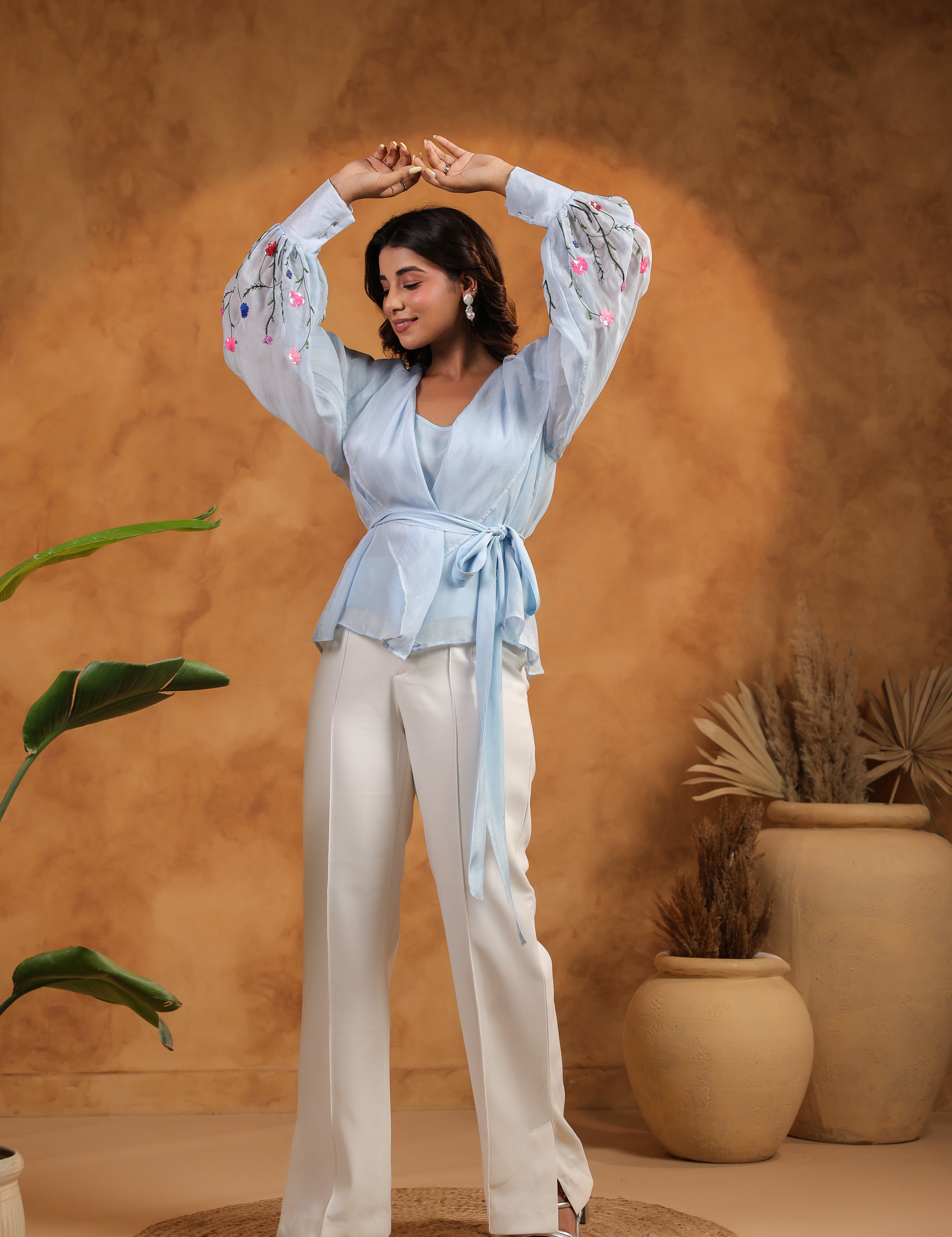 Woman in a light blue top and white pants posing against a warm brown background with plants.