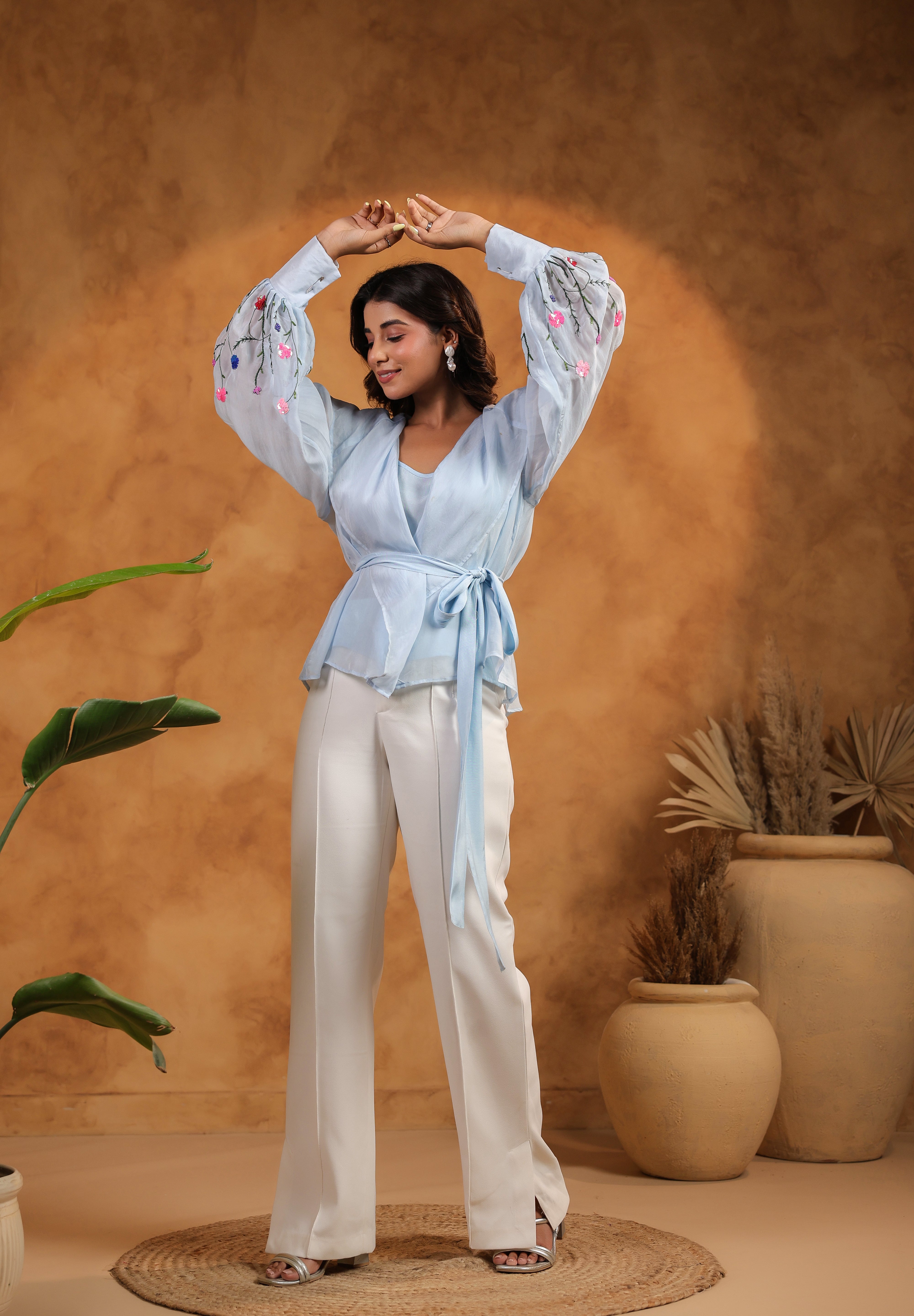 Woman in a light blue top and white pants posing against a warm brown background with plants.