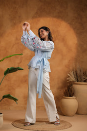 Woman in a light blue blouse and white pants standing against a warm brown background with plants.