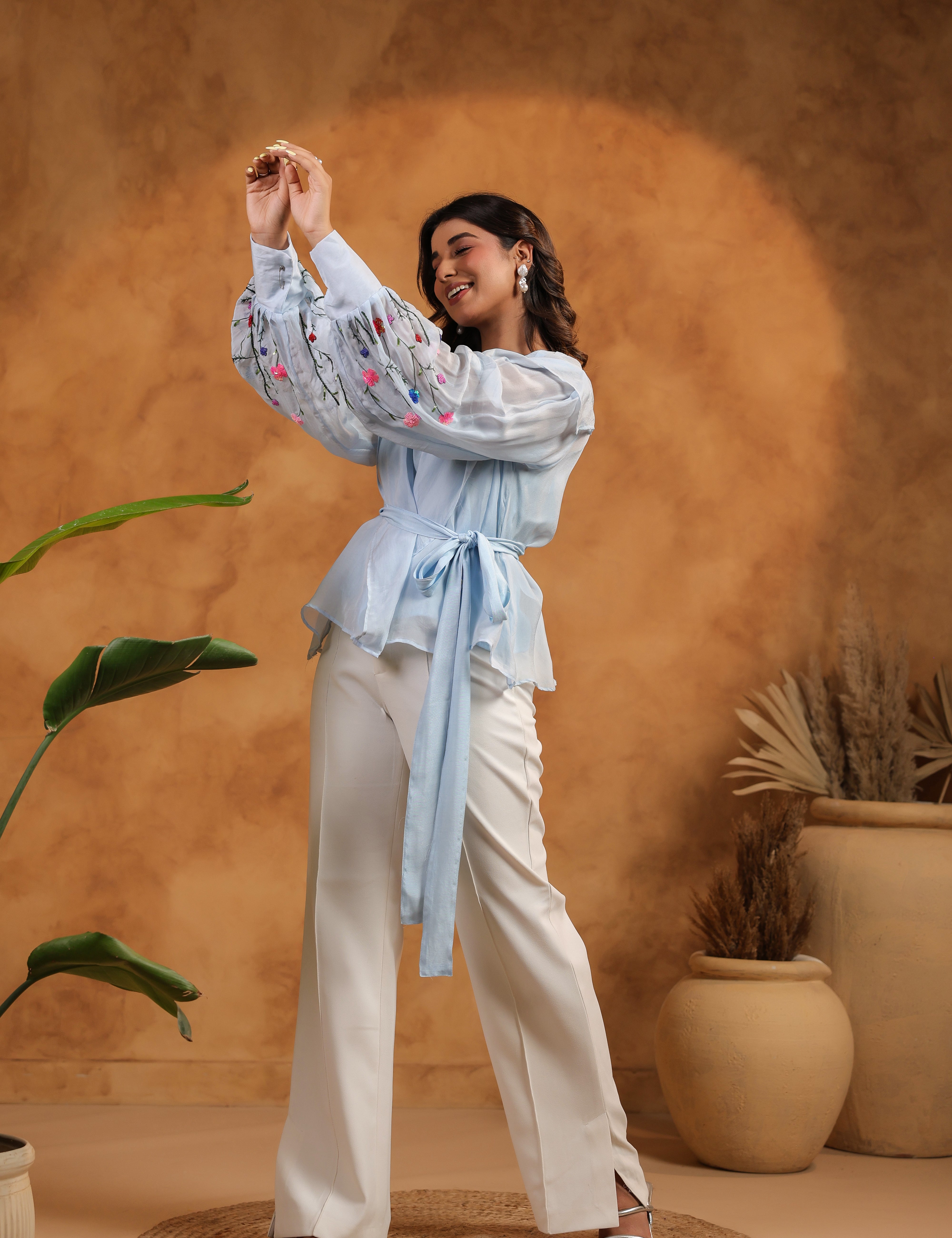 Woman in a light blue blouse and white pants standing against a warm brown background with plants.