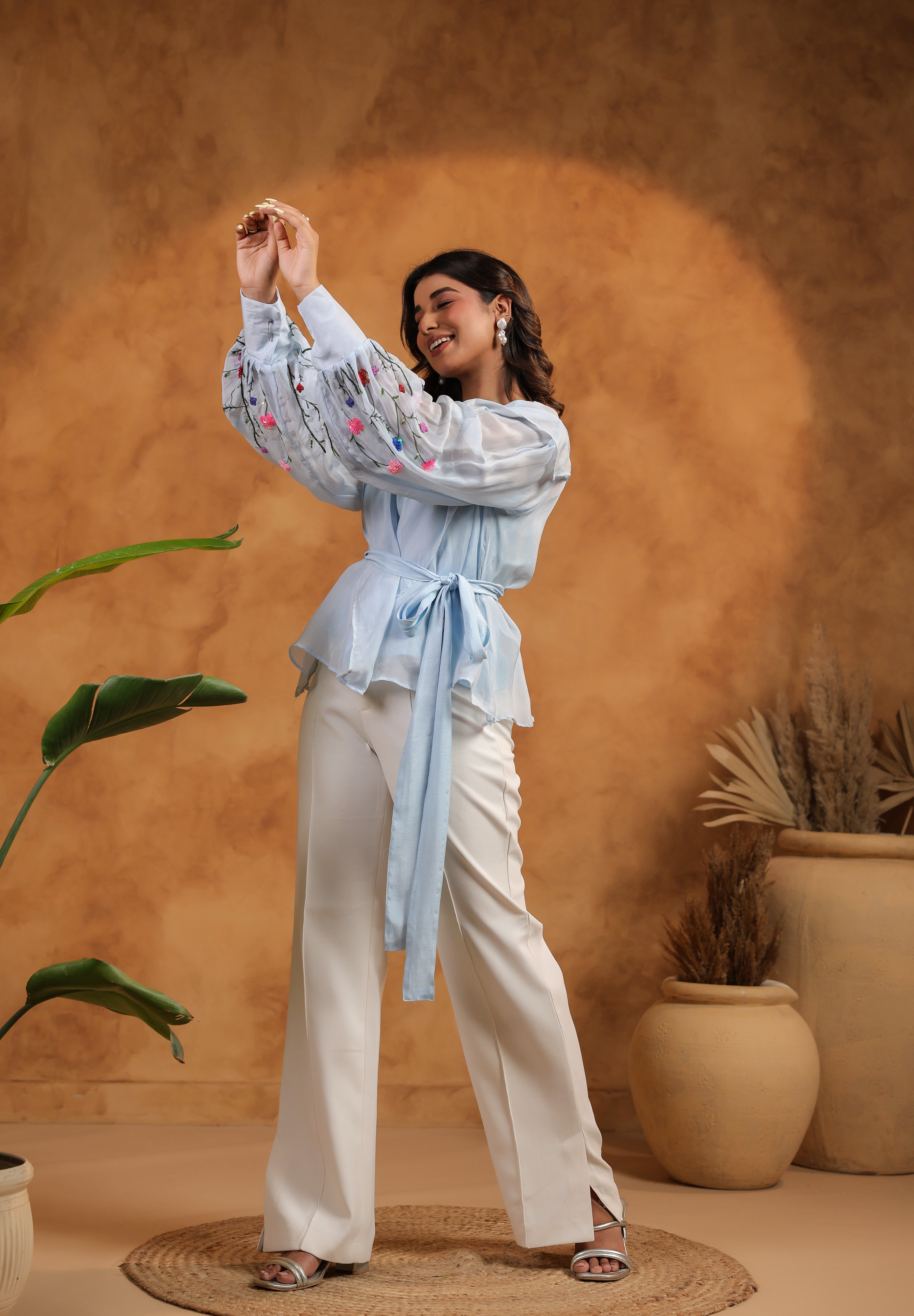 Woman in a light blue blouse and white pants standing against a warm brown background with plants.