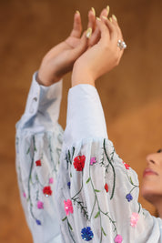 Person wearing a blue top with floral embroidery, raising hands against a blurred background