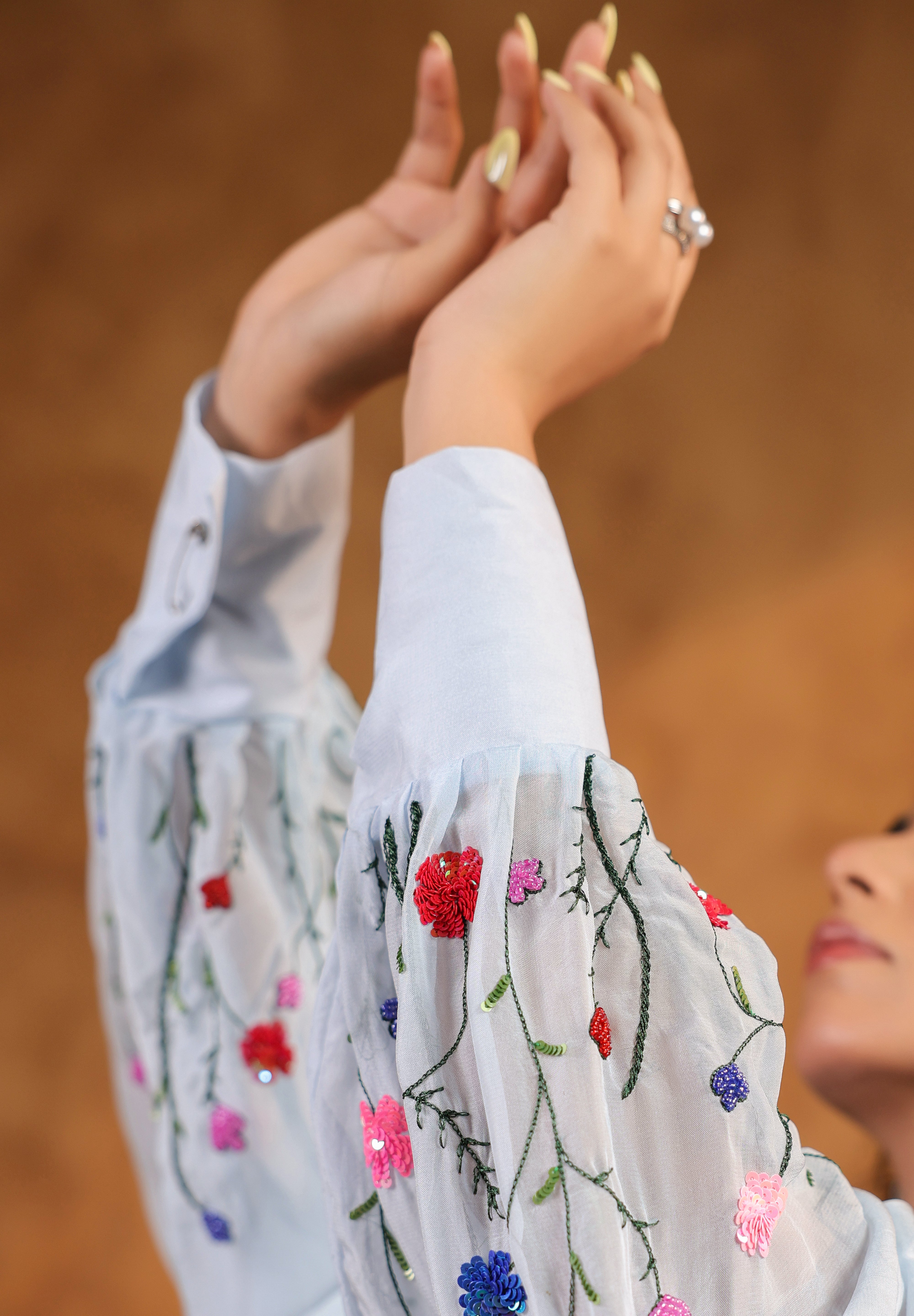 Person wearing a blue top with floral embroidery, raising hands against a blurred background