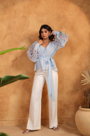 Woman wearing a light blue blouse with floral embroidery and white pants against a brown wall.