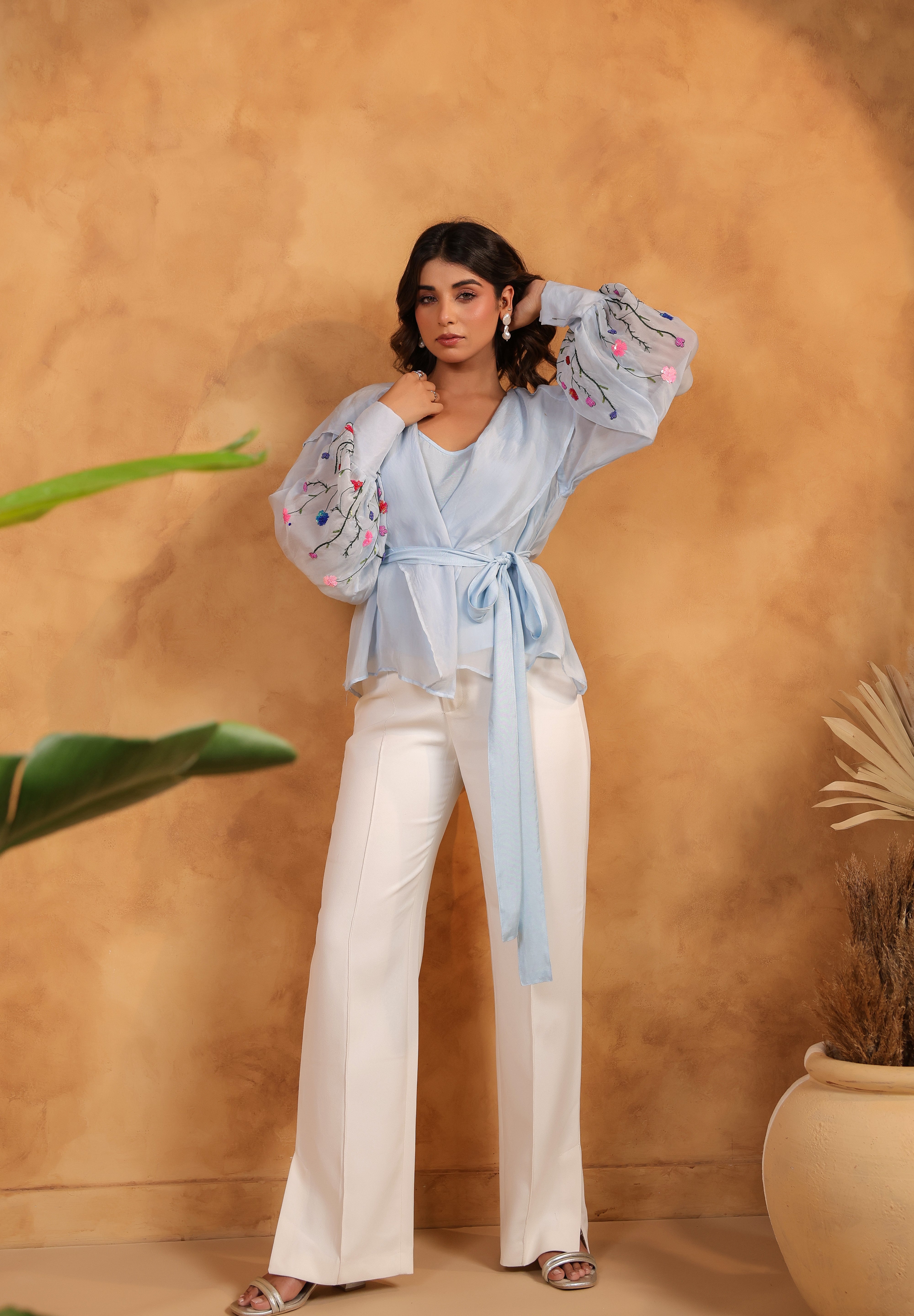 Woman wearing a light blue blouse with floral embroidery and white pants against a brown wall.