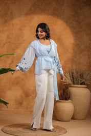 Woman in light blue top and white pants standing against a warm brown background with plants.