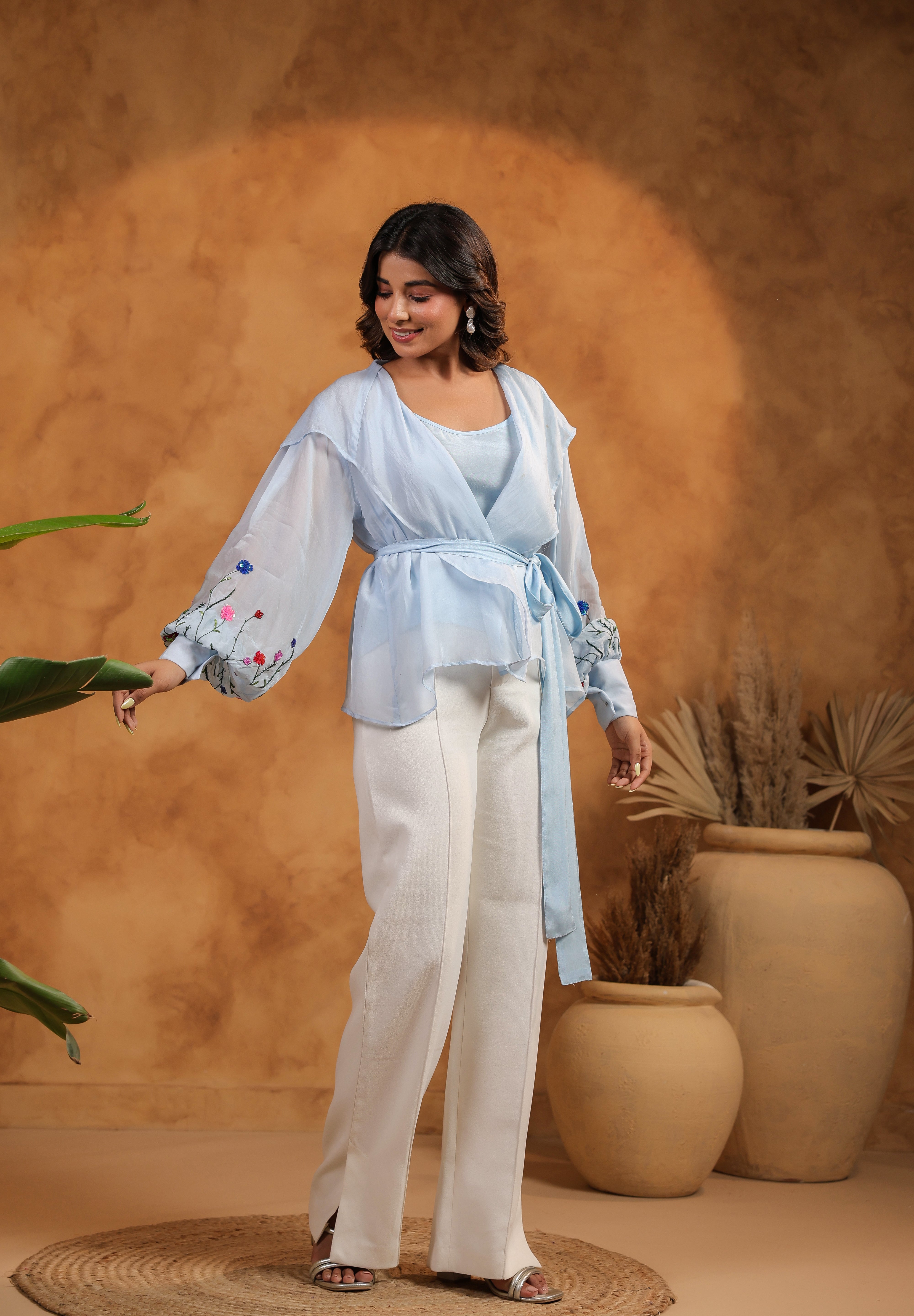Woman in light blue top and white pants standing against a warm brown background with plants.