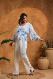Woman in a light blue top and white pants standing against a warm-toned wall with plants.