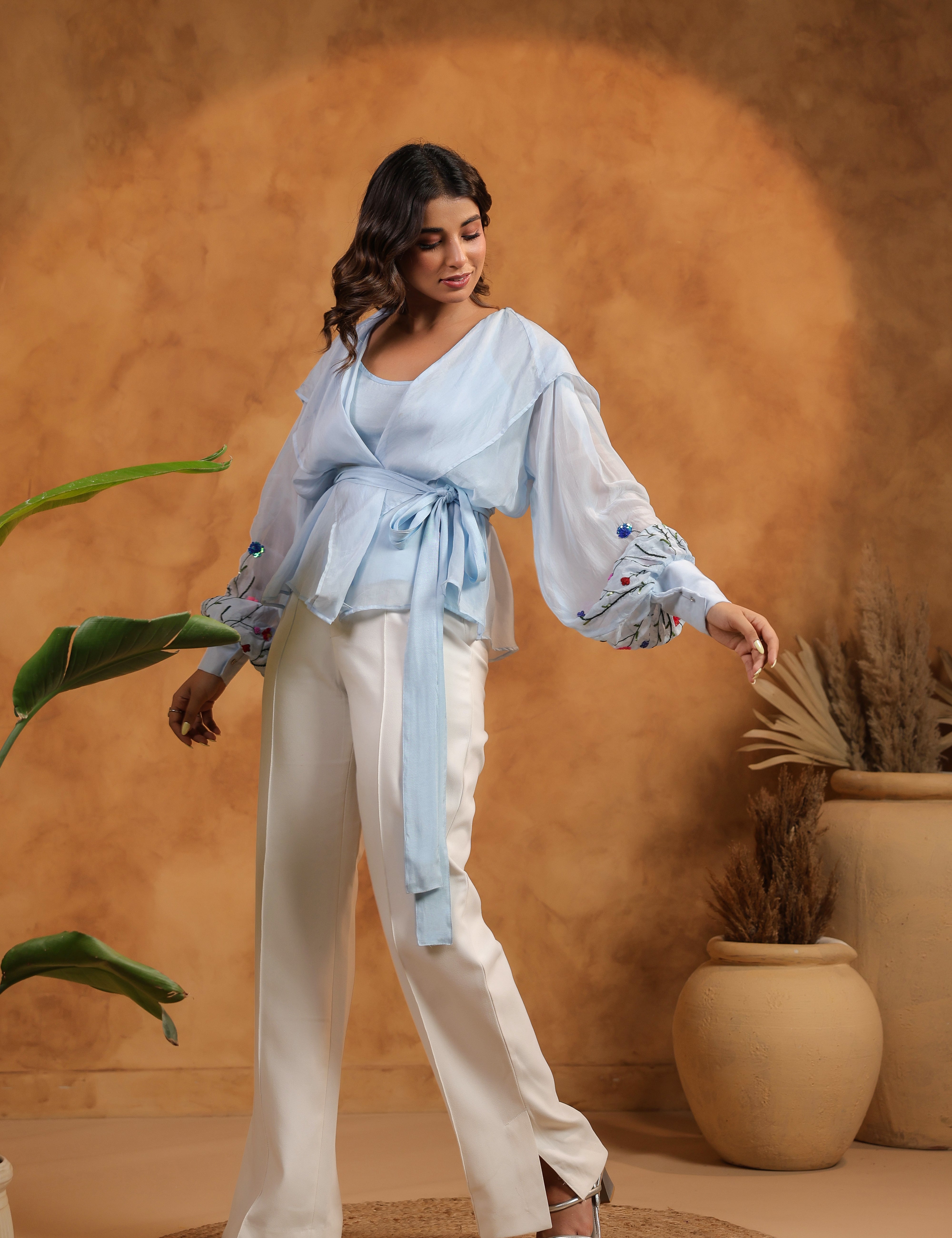 Woman in a light blue top and white pants standing against a warm-toned wall with plants.