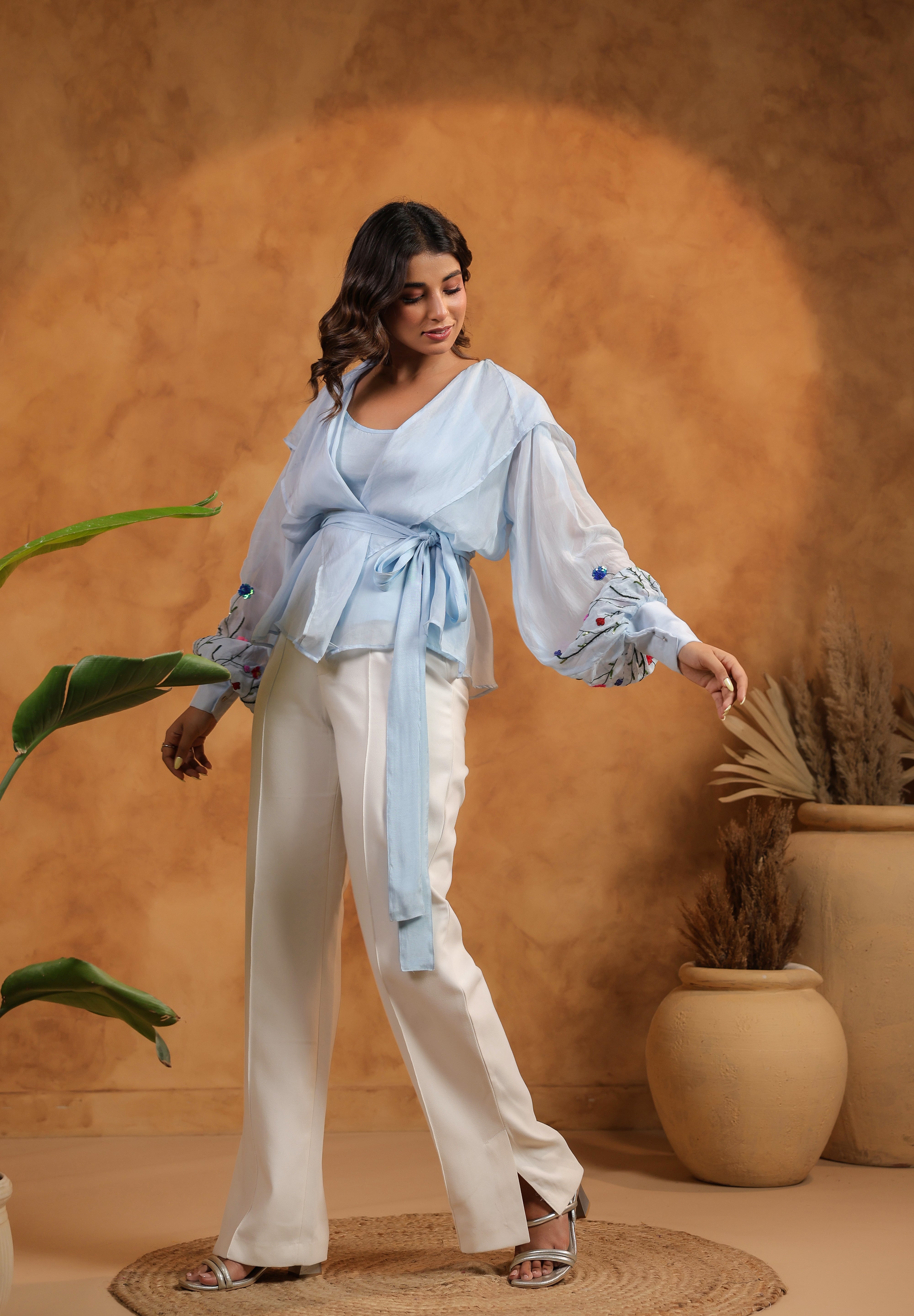Woman in a light blue top and white pants standing against a warm-toned wall with plants.