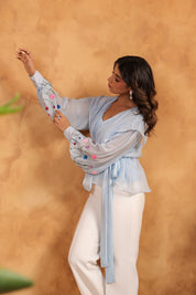 Woman wearing a light blue top with colorful pom-poms and white pants against a beige background